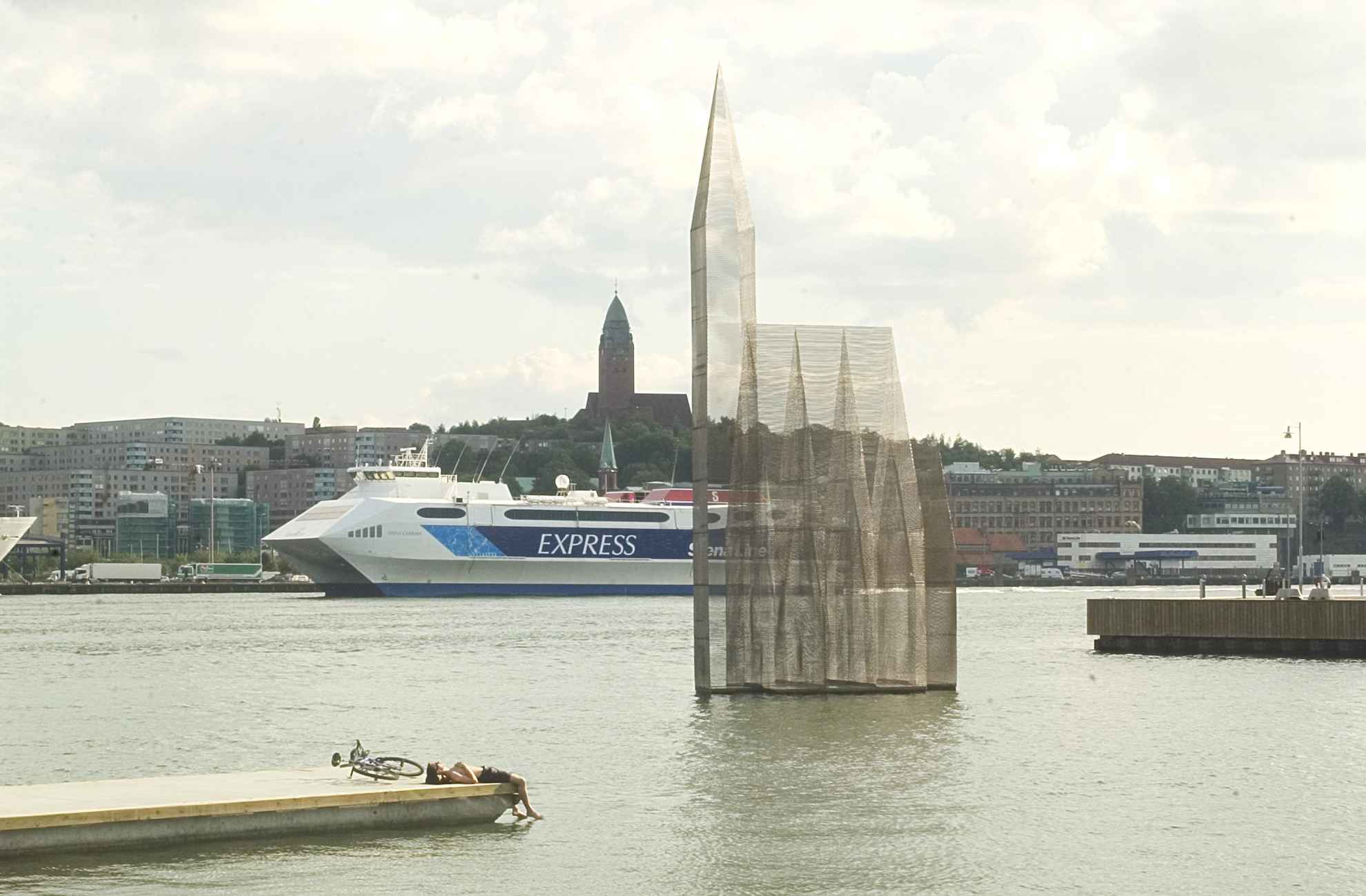 A transparent church-like sculpture called ‘Temple of Doubt and Hope’ floats on the water in in Gothenburg harbour basin. To the left a person is lying on a jetty next to a bike. In the background you see a ferryboat.