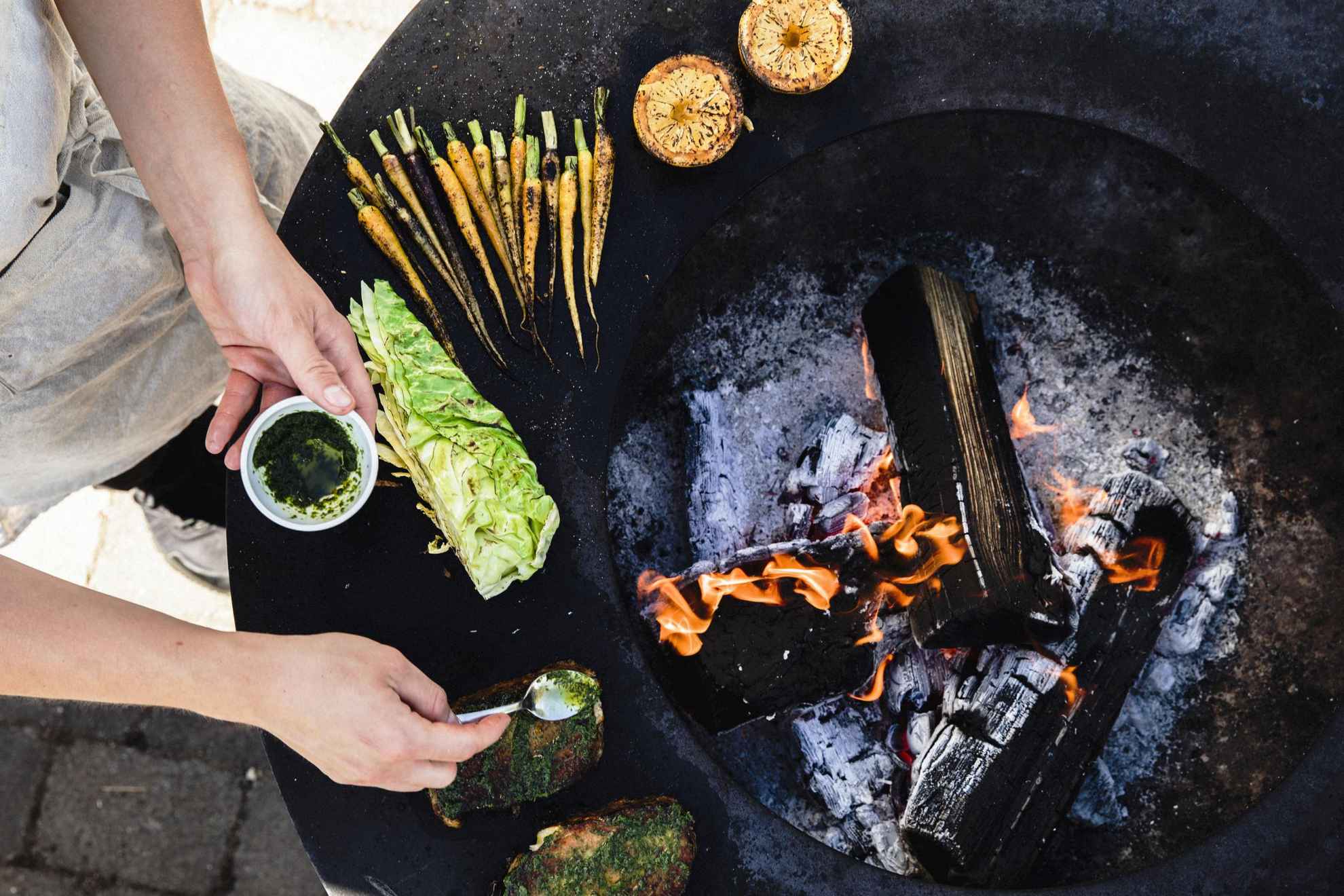 Un chef prépare de la nourriture à côté d'un feu. A côté du feu, il y a des champignons, des carottes, de la salade et du pain.