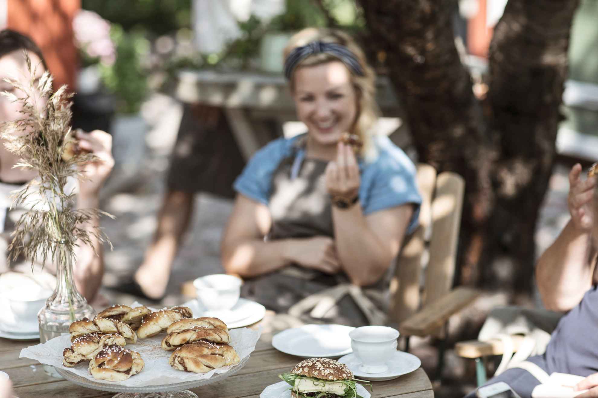 Une femme assise à une table en plein air mange une brioche à la cannelle. Sur les tables se trouvent une assiette de brioches à la cannelle, un sandwich et deux tasses à café.