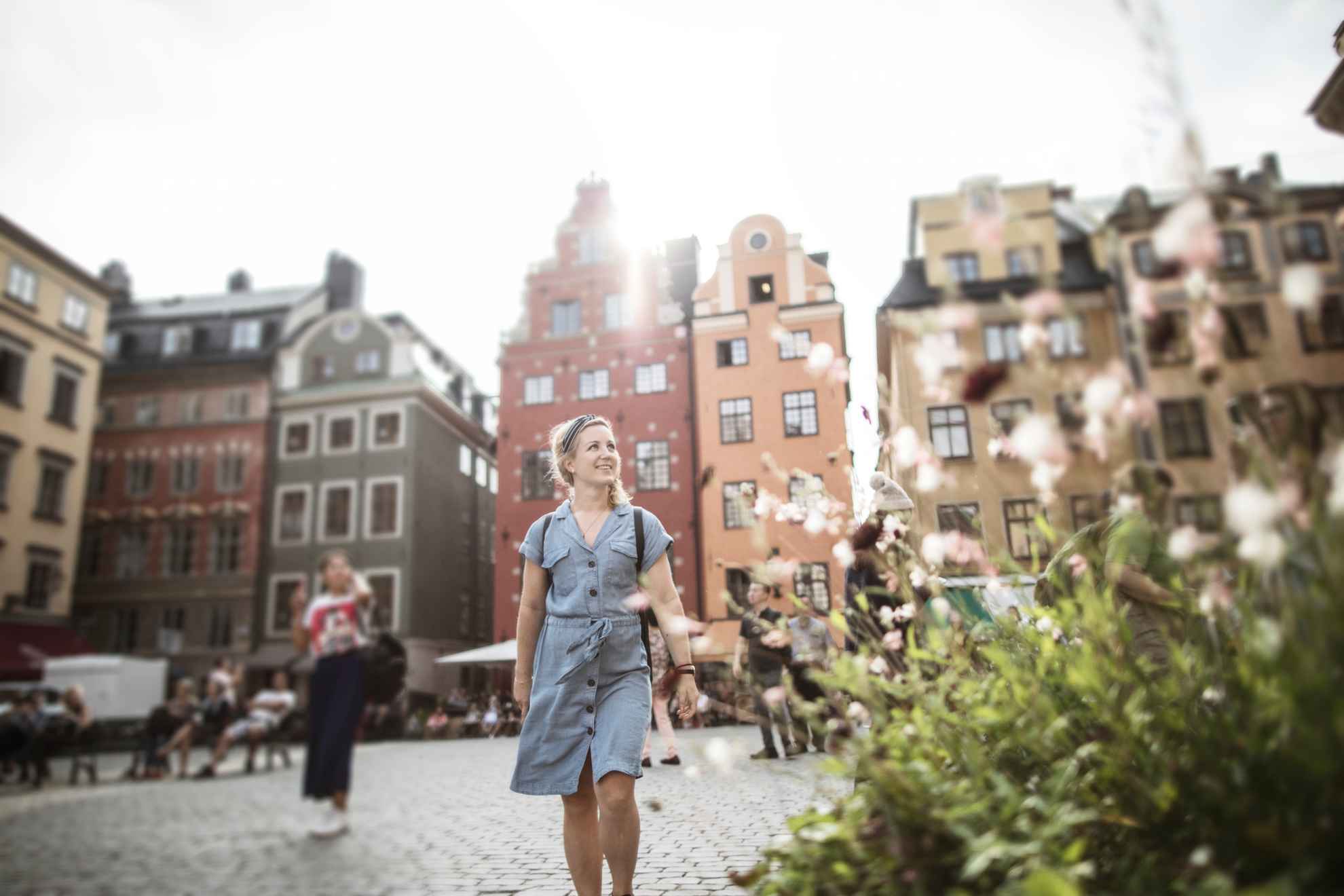 Une femme marchant sur une place de la vieille ville. À l'arrière-plan, on aperçoit de vieilles maisons en pierre et, à l'avant, se trouve une fleur.