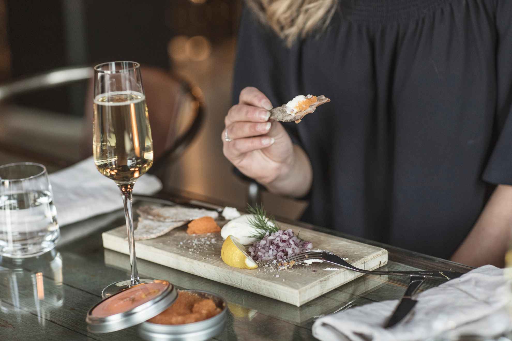 Gros plan sur une femme mangeant du caviar, de l'oignon rouge, de la crème, du citron et du pain croustillant sur un plateau en bois et buvant un verre de mousseux.