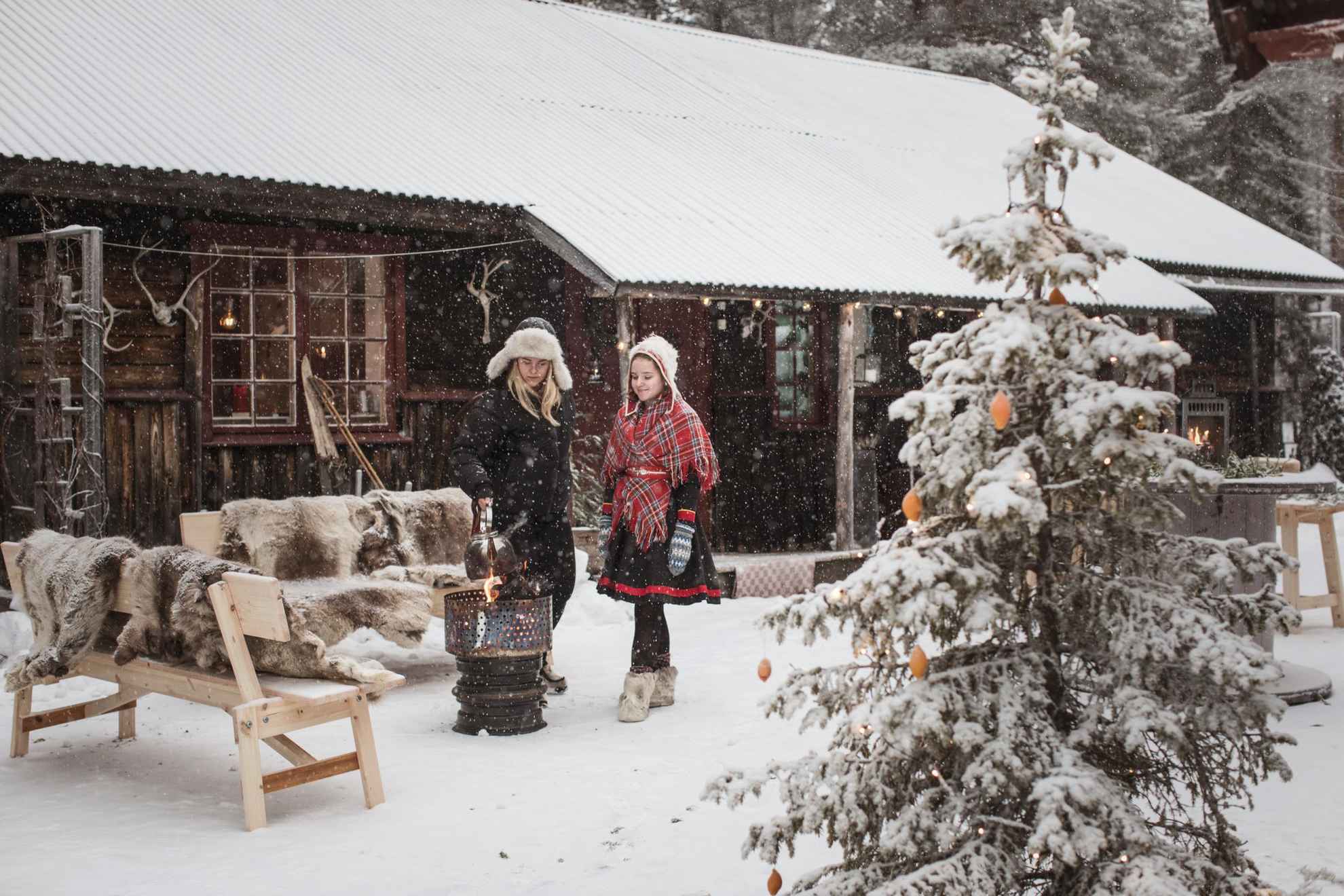 Deux jeunes femmes sont debout dans la neige devant une maison en bois. L'une des filles porte une tenue samie. Elles cuisinent sur un feu ouvert. Il y a des fourrures de rennes sur les bancs et un sapin au premier plan.