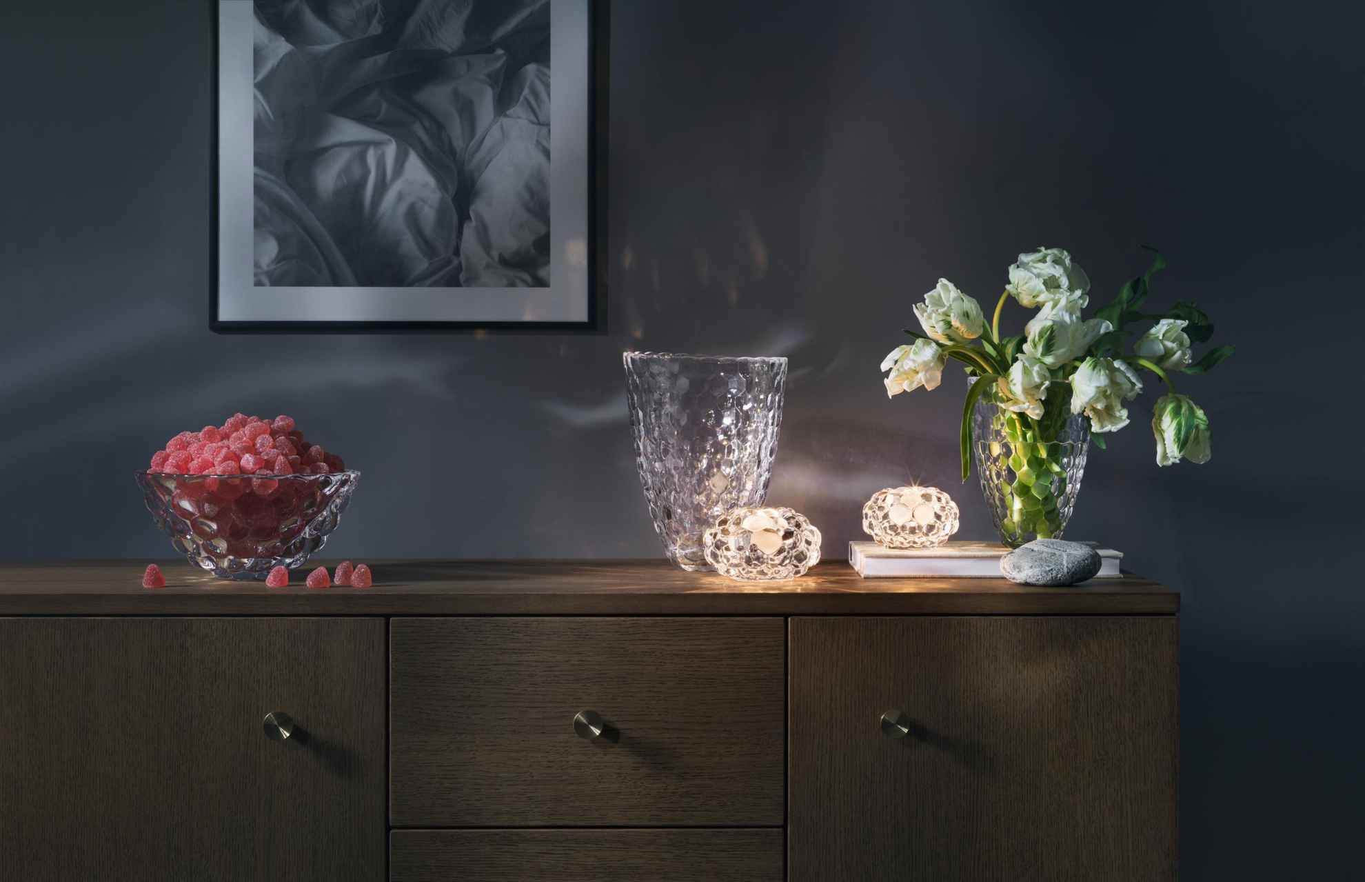 Objets design et en verre posés sur un meuble en bois. Sur ce meuble sont disposés deux bougeoirs avec des bougies allumées et deux vases, l'un avec des tulipes et l'autre rempli de bonbons rouges.