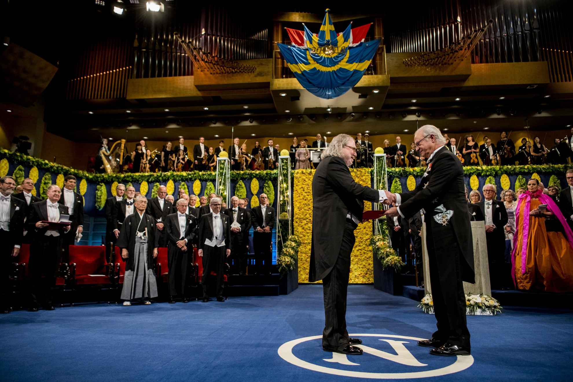 Un lauréat masculin est debout sur une scène et reçoit son prix des mains du roi de Suède, Carl XVI Gustaf.