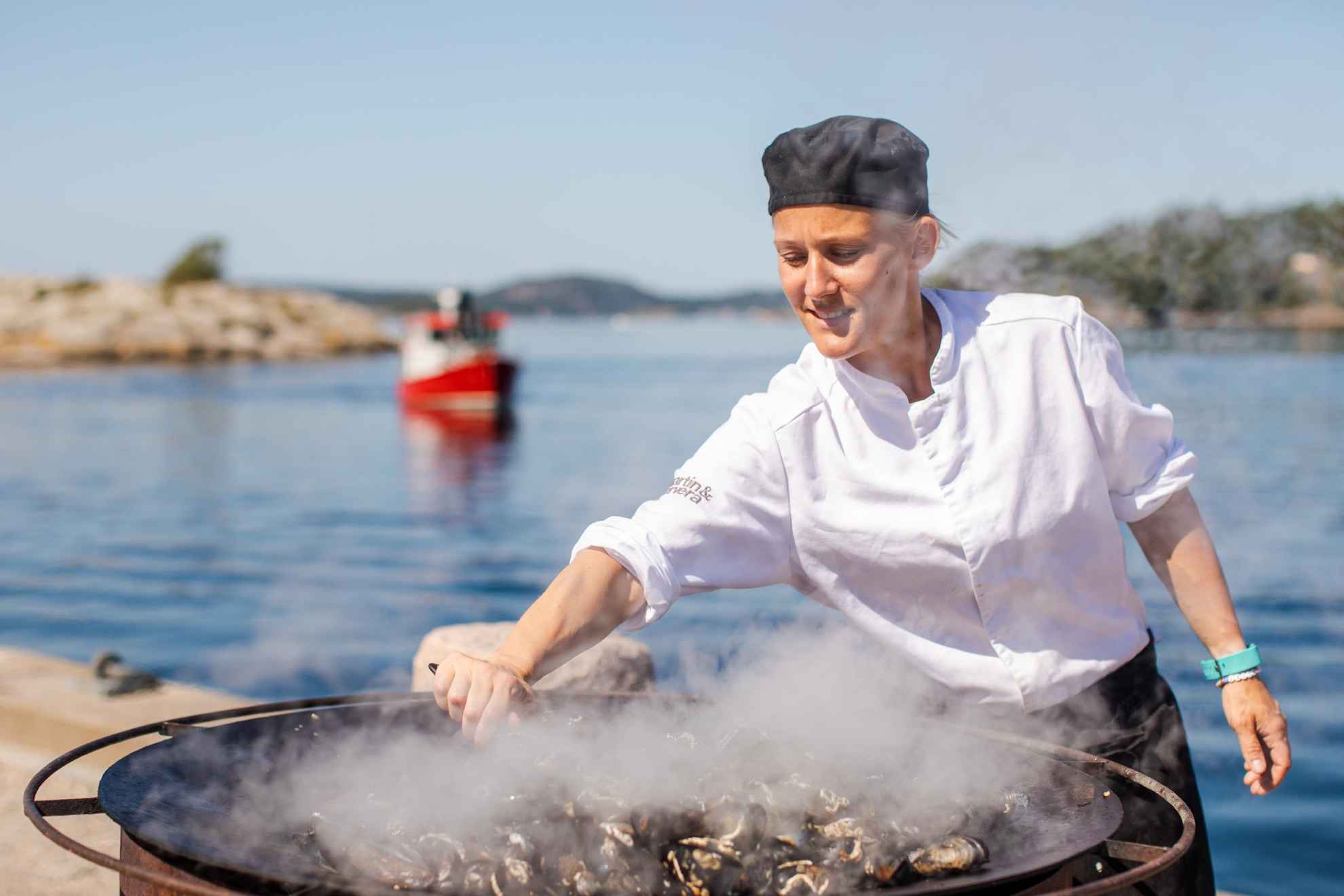 Un chef se tient dehors au bord de la mer et cuisine des moules dans une grande poële.