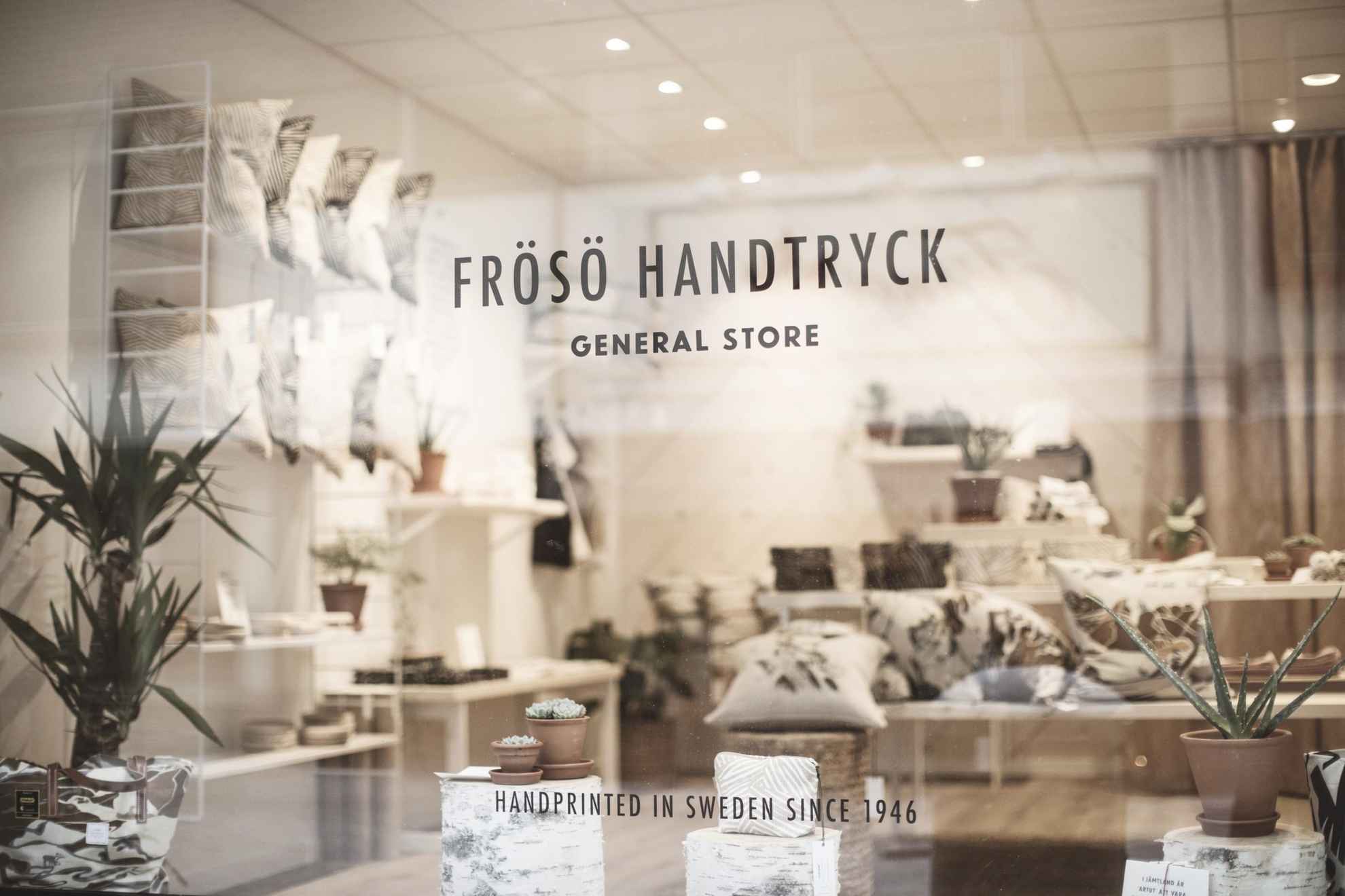 Vue sur un magasin de design d'intérieur depuis la vitrine. Il y a des textiles et des oreillers. Un logo collé sur la fenêtre indique Frösö Handtryck.
