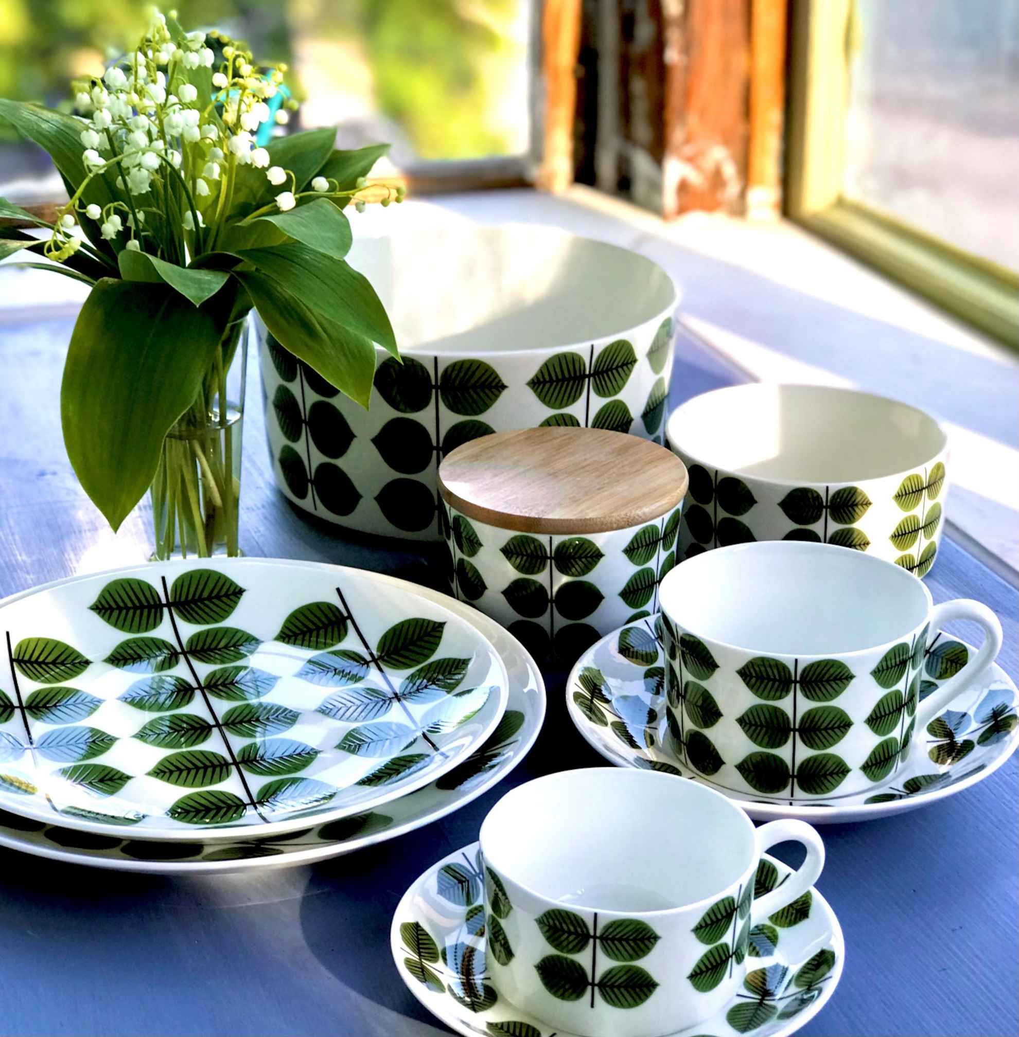 Gros plan sur un ensemble de porcelaine posé sur une table et composé de vaisselle peinte de feuilles vertes et d’un vase avec des muguets.