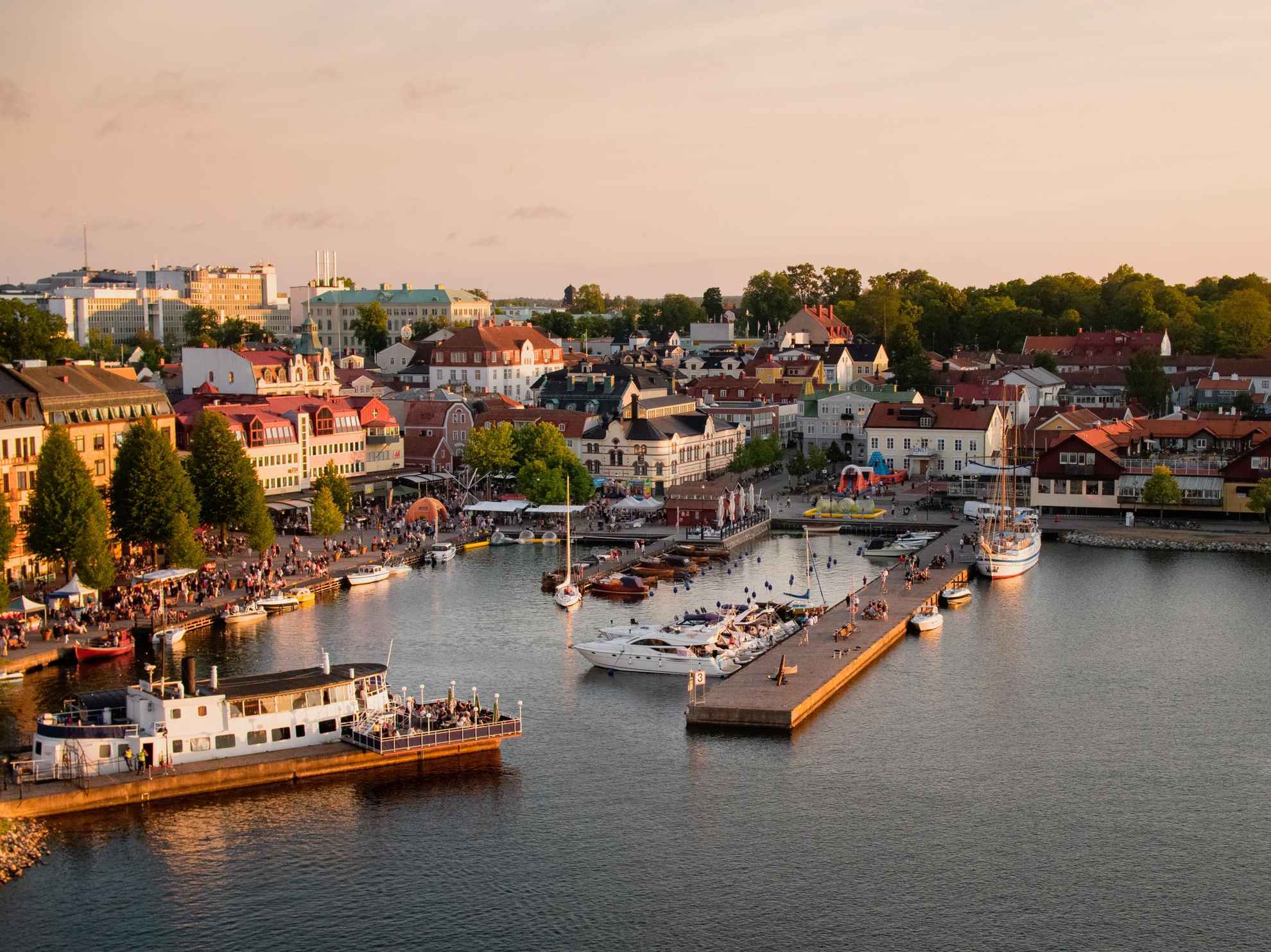 Vue sur le centre-ville de Västervik avec son port. Plusieurs bateaux sont amarrés aux quais. Il y a beaucoup de monde rassemblé sur la jetée et dans les terrasses des restaurants.