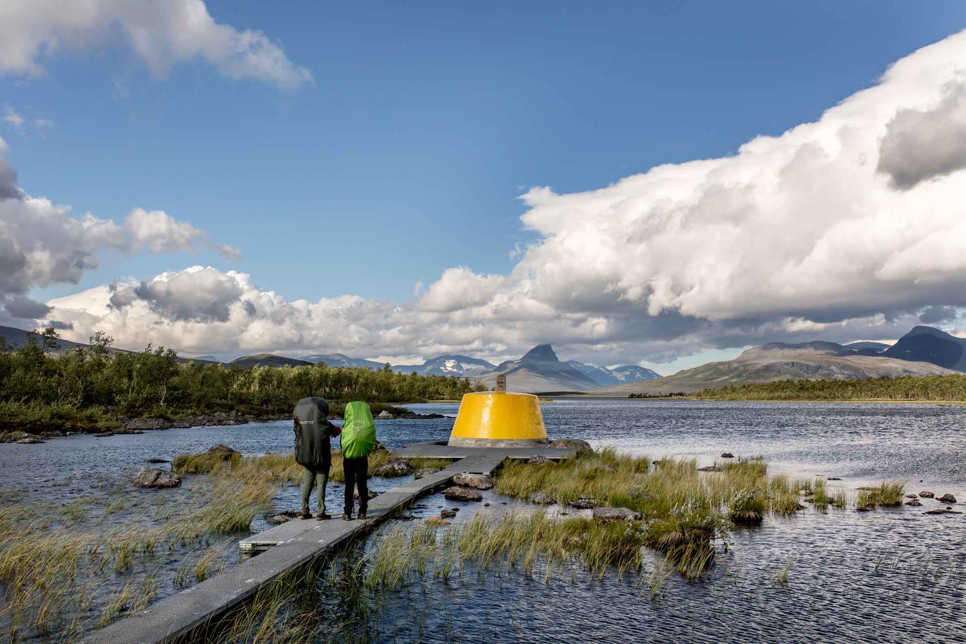 Deux randonneurs avec des sacs à dos regardent un grand cairn en béton dans l'eau avec les montagnes en arrière-plan.