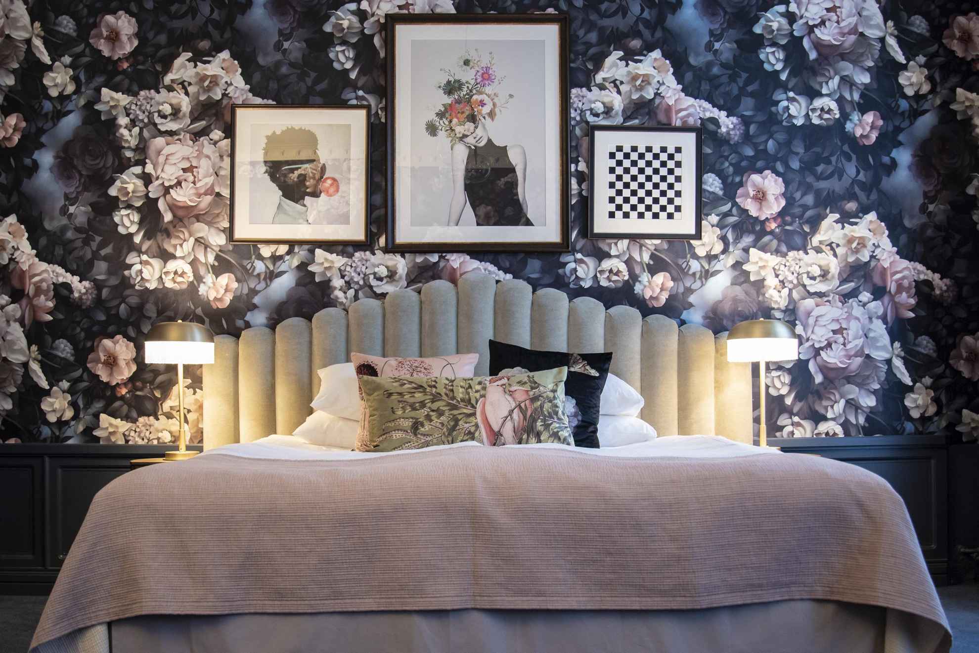 Vue sur l'intérieur d'une chambre d'hôtel. On voit un lit devant un mur recouvert de papier peint avec de grandes roses rose et blanches. Sur le mur sont accrochés trois tableaux et quatre coussins aux motifs différents sont disposés sur le haut du lit.