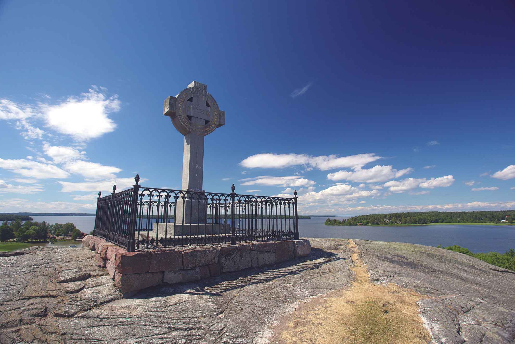 La croix d'Ansgar se dresse au sommet d'une falaise. On aperçoit en arrière-plan, le lac Mälaren, des îles et un ciel bleu.
