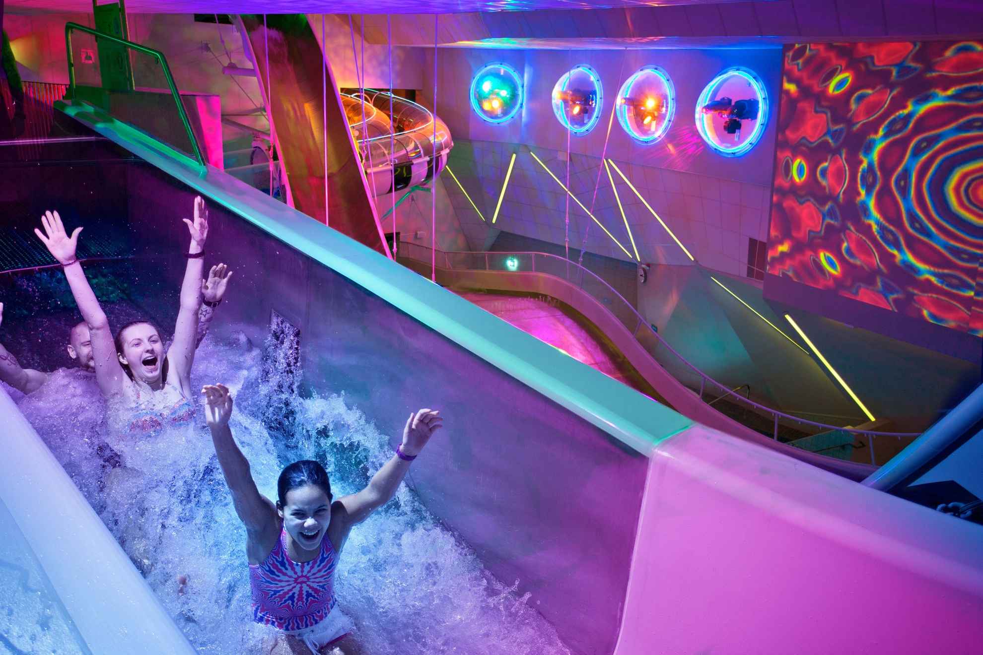 Trois personnes sourient et lèvent les bras en descendant un toboggan aquatique. L'endroit est principalement éclairé par des lumières violettes et des néons.