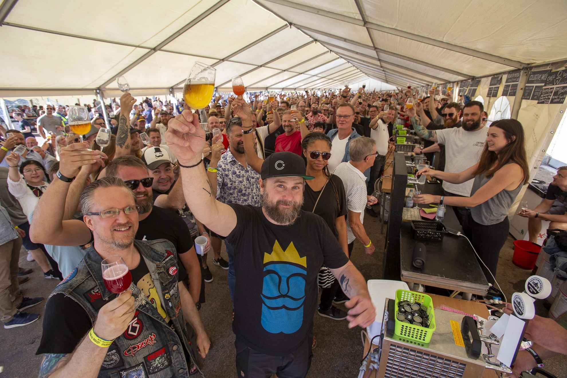 Une tente au festival de Brewskival remplie de personnes en train de porter un toast avec leur bière en direction de la caméra.