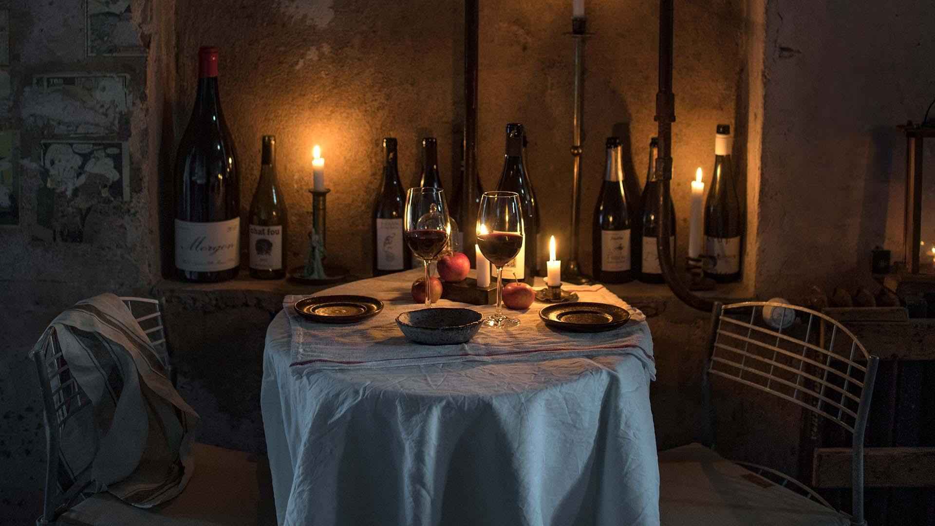 Une table pour deux personnes avec une nappe blanche, des assiettes et deux verres remplis de vin rouge. Le mur est décoré avec des bouteilles de vin vides et des bougies allumées.