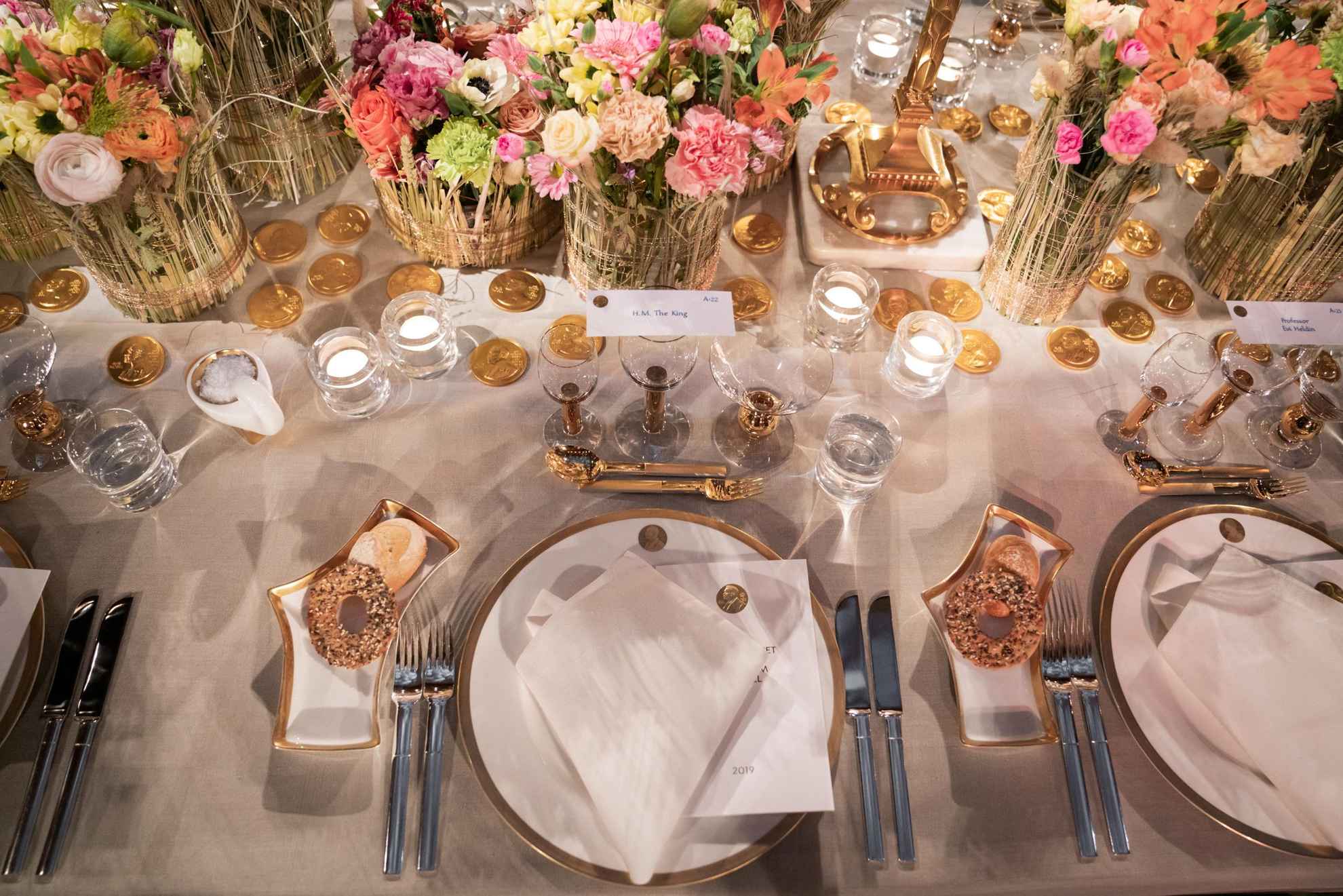 Une vaisselle blanche avec des détails dorés est posée sur une table avec une nappe blanche. Au milieu de la table se trouvent des fleurs colorées dans des vases.