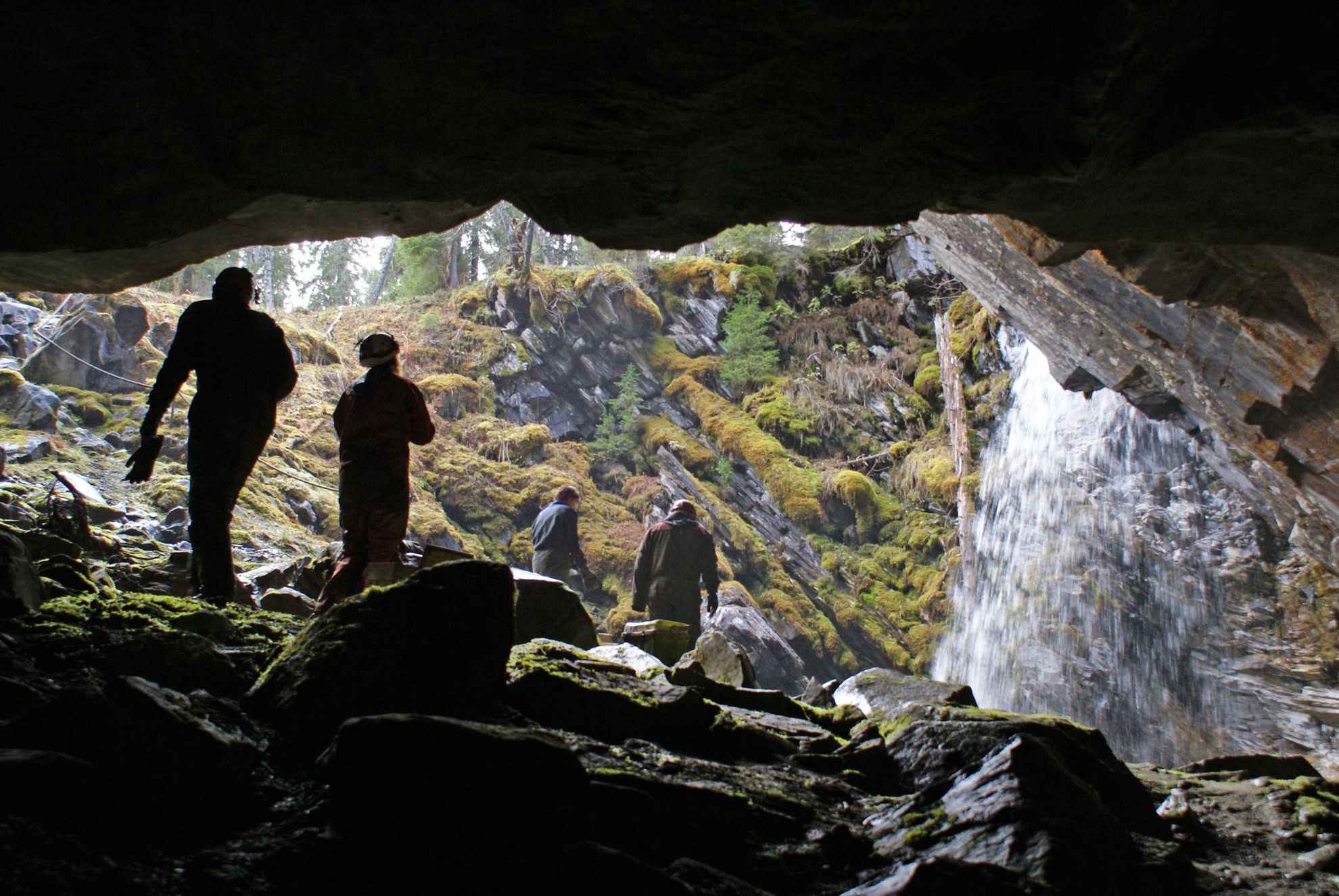 Vue de l'intérieur d'une grotte donnant sur une clairière avec quatre personnes marchant vers une cascade.