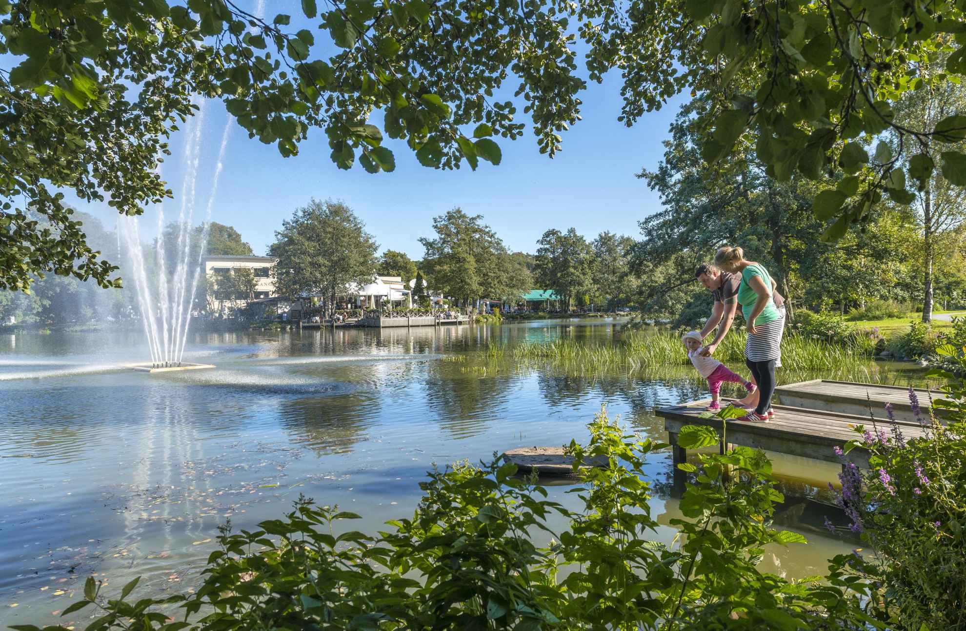 Un couple et un petit enfant sont debout sur un ponton à côté d'un étang avec une fontaine. Ils sont dans le parc de Slottsskogen et sont entourés de verdure. En arrière-plan, on peut voir un restaurant avec une partie extérieure.