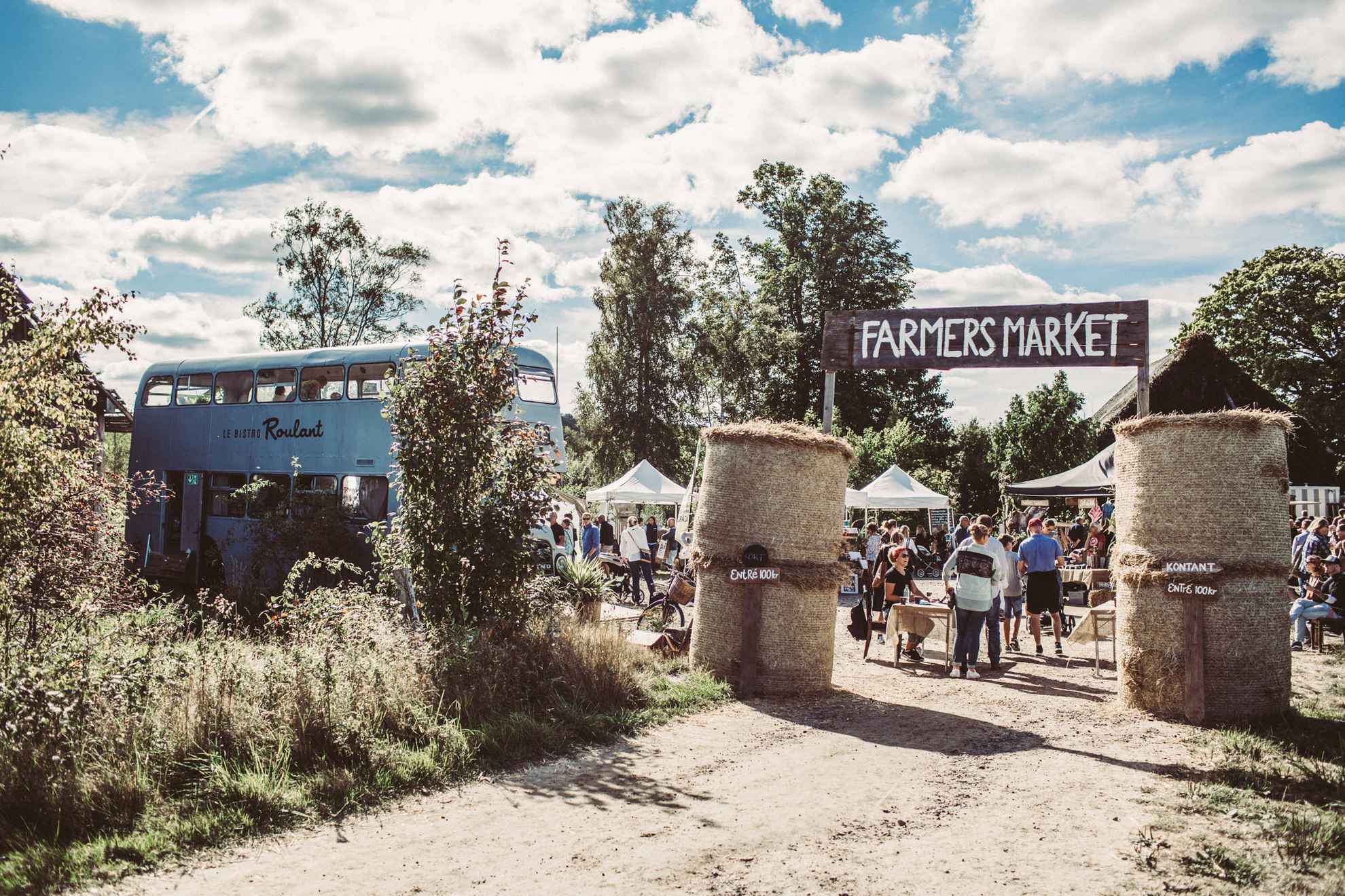 On peut voir un grand panneau indiquant « Farmers Market » à l'entrée d'un marché rural. Un bus bleu clair est garé à côté.