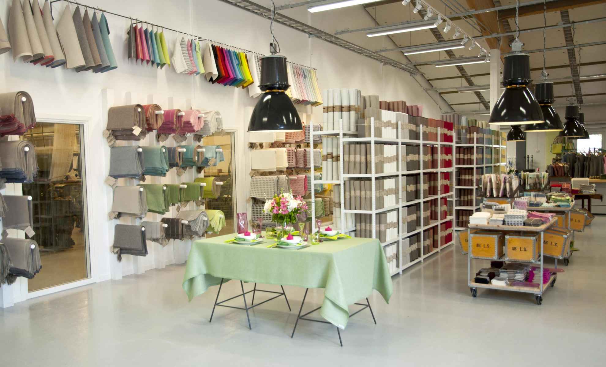 Intérieur d'un magasin d'usine avec des produits en lin. Les étagères et les tables sont remplis de textiles et produits en lin. Une table est dressée avec une nappe verte et des serviettes.