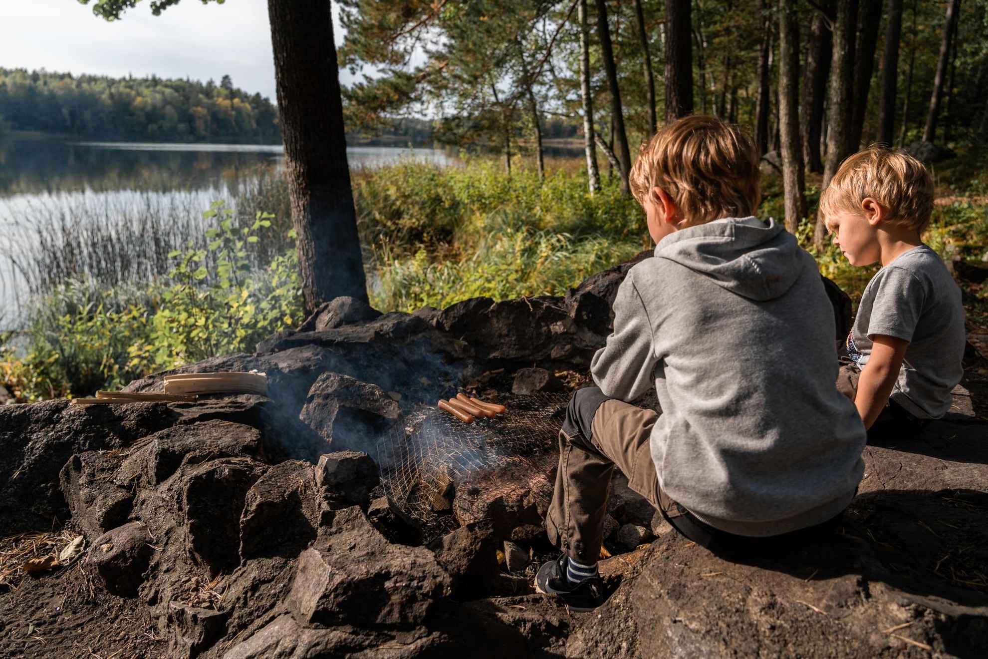 Deux enfants sont assis dehors dans la nature à côté d'un feu et grillent des saucisses.