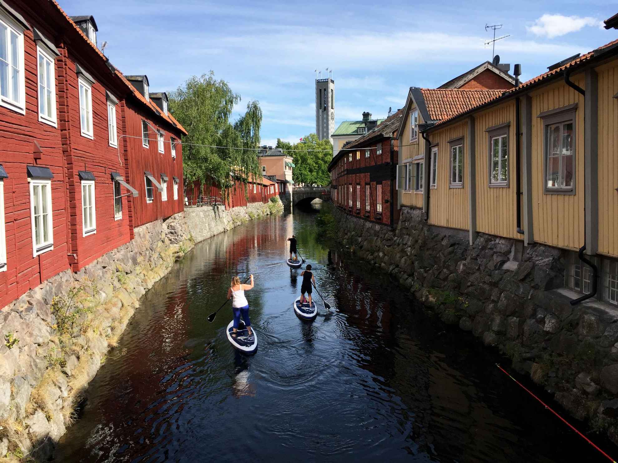Trois personnes pagayent sur des planches de stand up paddle sur un canal avec des maisons de chaque côté.