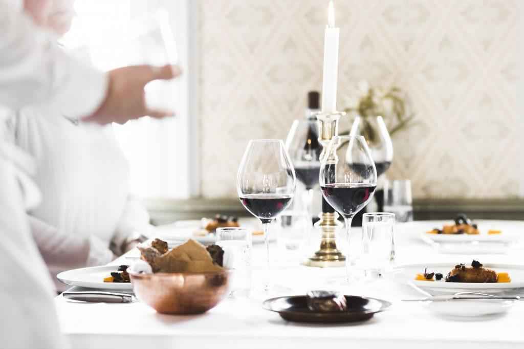 Une table avec une nappe blanche, une bougie allumée, des assiettes avec de la nourriture et des verres remplis de vin rouge. Un serveur est au premier plan de la photo.