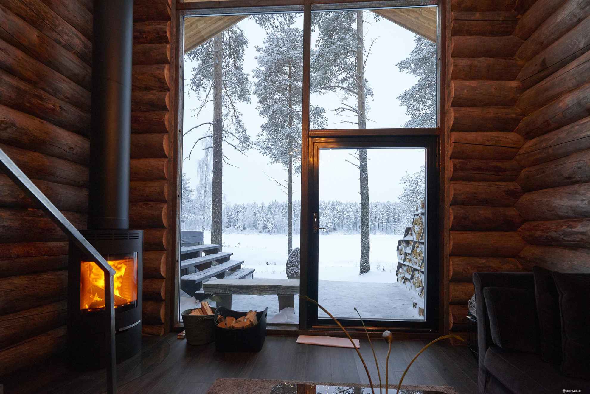 Une cabane en rondins en bois comprenant de larges ouvertures et un feu de cheminée. Il neige dehors.
