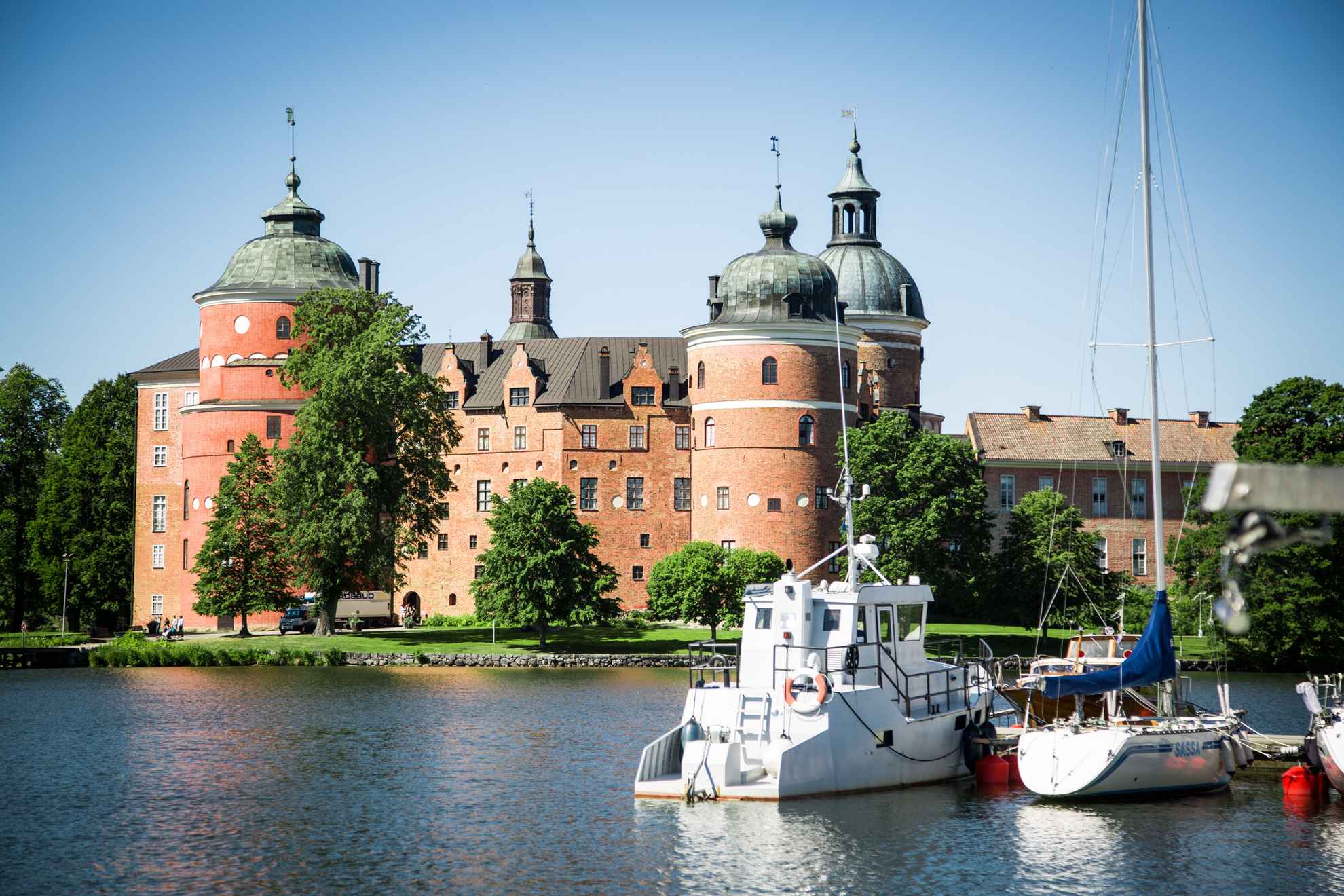 Un château entouré de verdure se trouve de l'autre côté d'un lac. Le soleil brille et des bateaux sont amarrés à l'embarcadère.