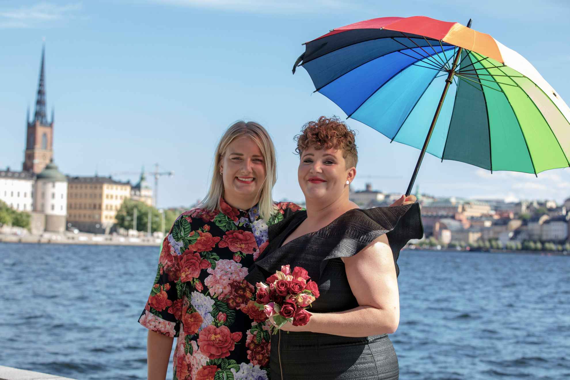 Deux femmes, l'une avec une robe noire et l'autre avec une chemise à fleurs, tiennent un parapluie aux couleurs de l'arc-en-ciel. Elles se tiennent au bord de l'eau, avec une vue de la ville de Stockholm en arrière-plan.