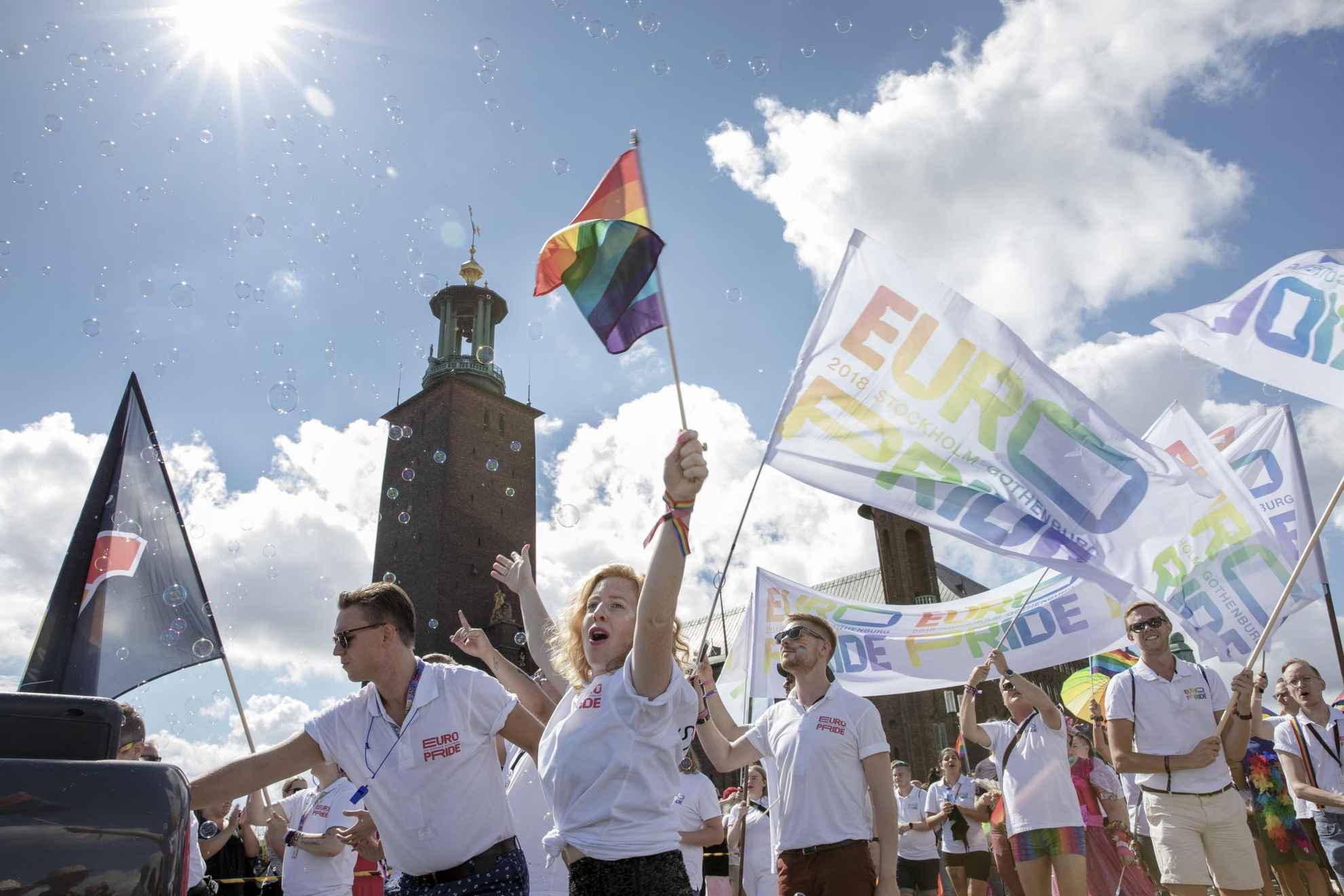 Plusieurs personnes marchant dans un défilé de la fierté. Ils ont des drapeaux arc-en-ciel et des drapeaux avec le texte Euro Pride. L'Hôtel de ville de Stockholm est en arrière-plan.
