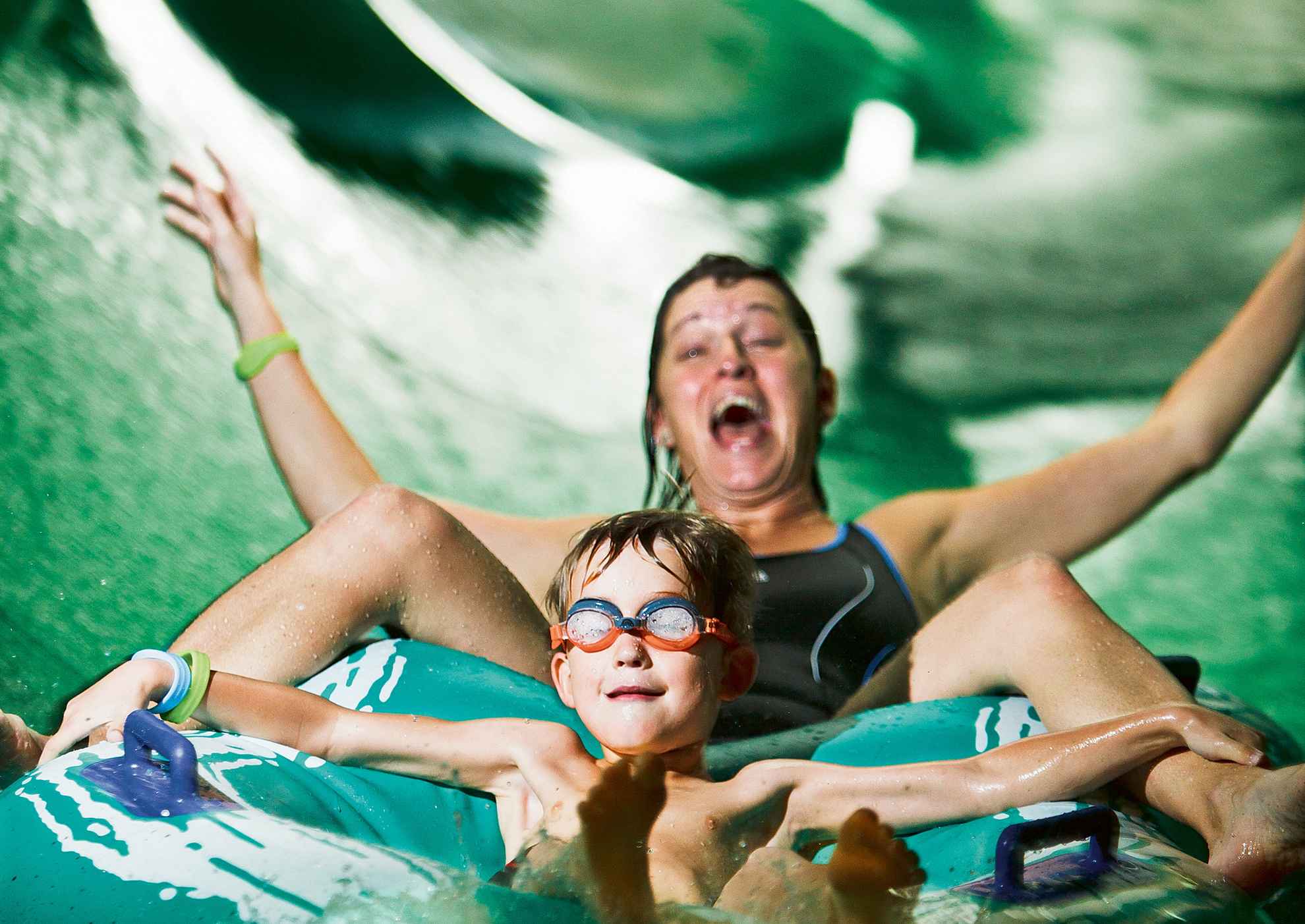 Un garçon souriant avec des lunettes de natation sur un toboggan aquatique avec une femme qui rit.