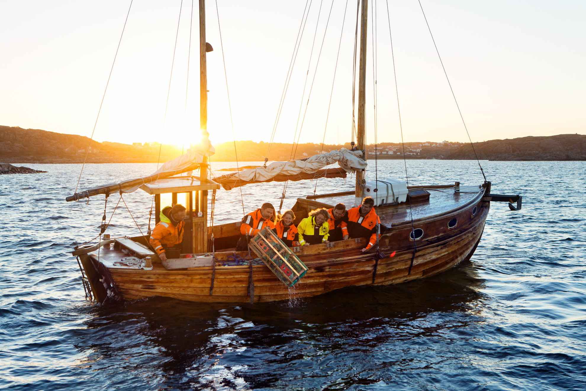 Un voilier en bois avec six personnes, dont l'une jette une cage à homard à la mer. Le soleil se couche en arrière-plan.