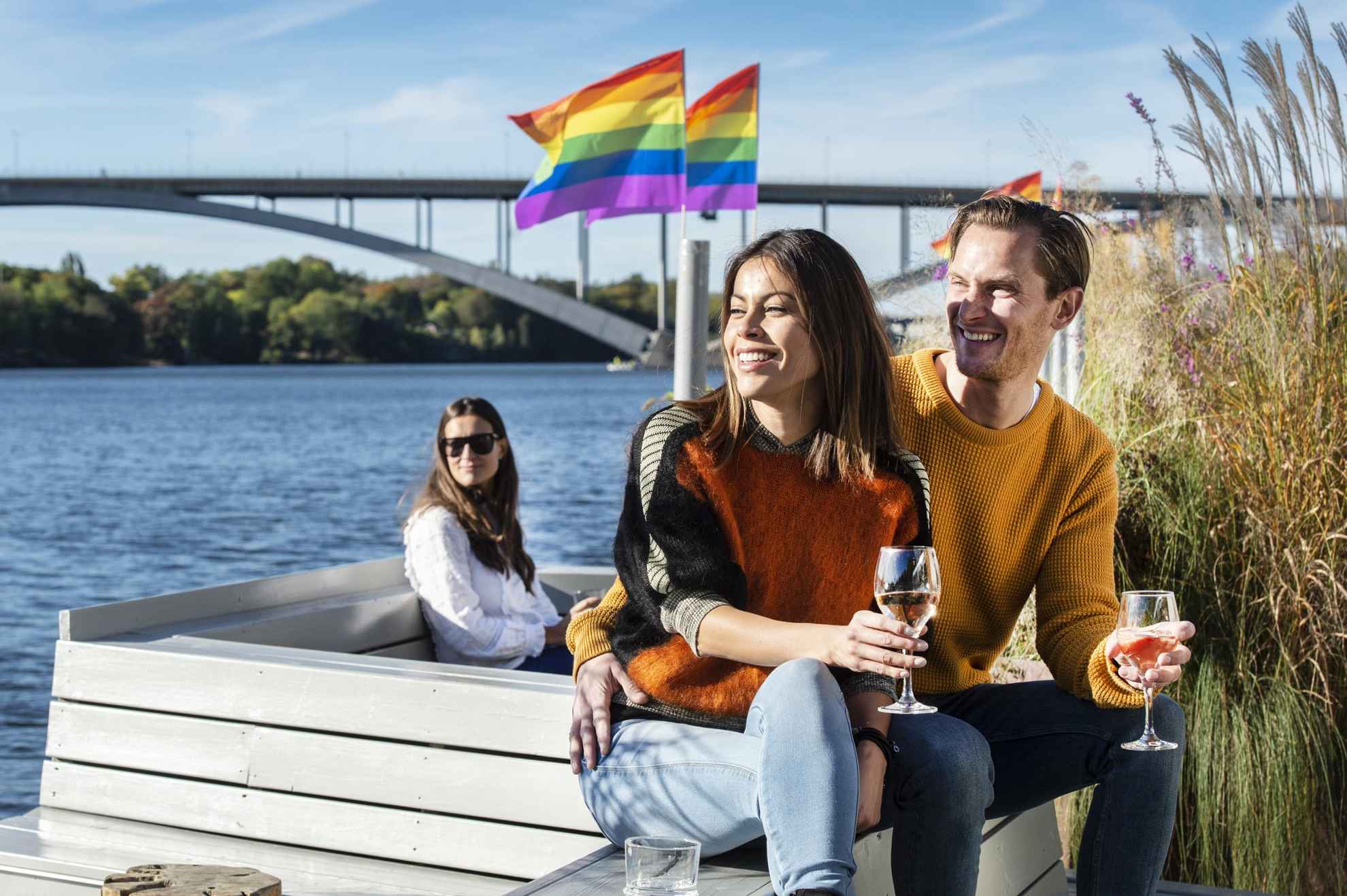 Un homme et une femme assis l'un à côté de l'autre sur une terrasse au bord de la mer, tenant chacun un verre de vin. Derrière eux, il y a une femme assise sous des drapeaux arc-en-ciel.