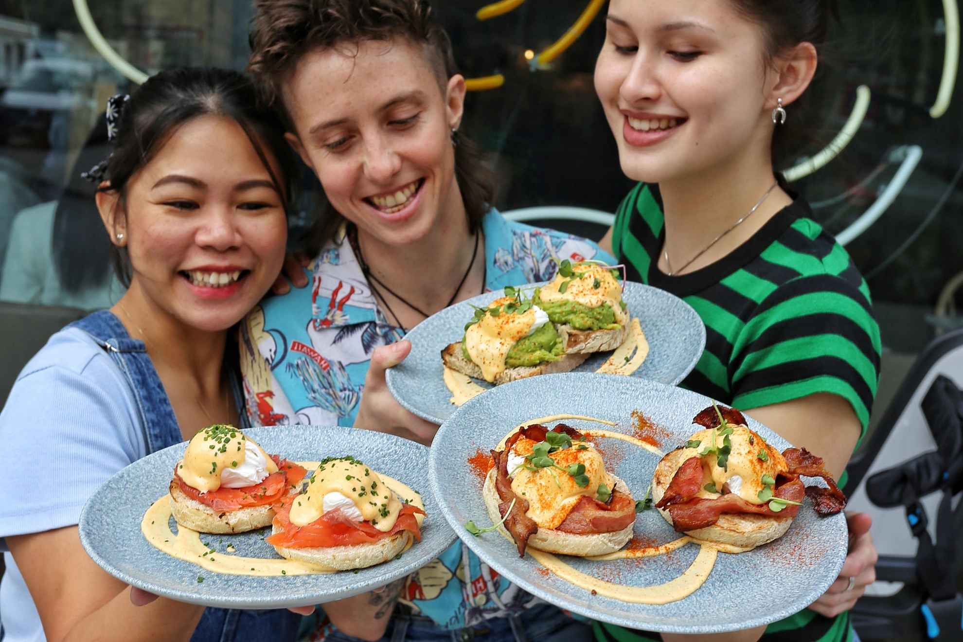 Trois femmes sourient et portent des assiettes contenant différents sandwichs. Elles se tiennent devant le restaurant Greasy Spoon.