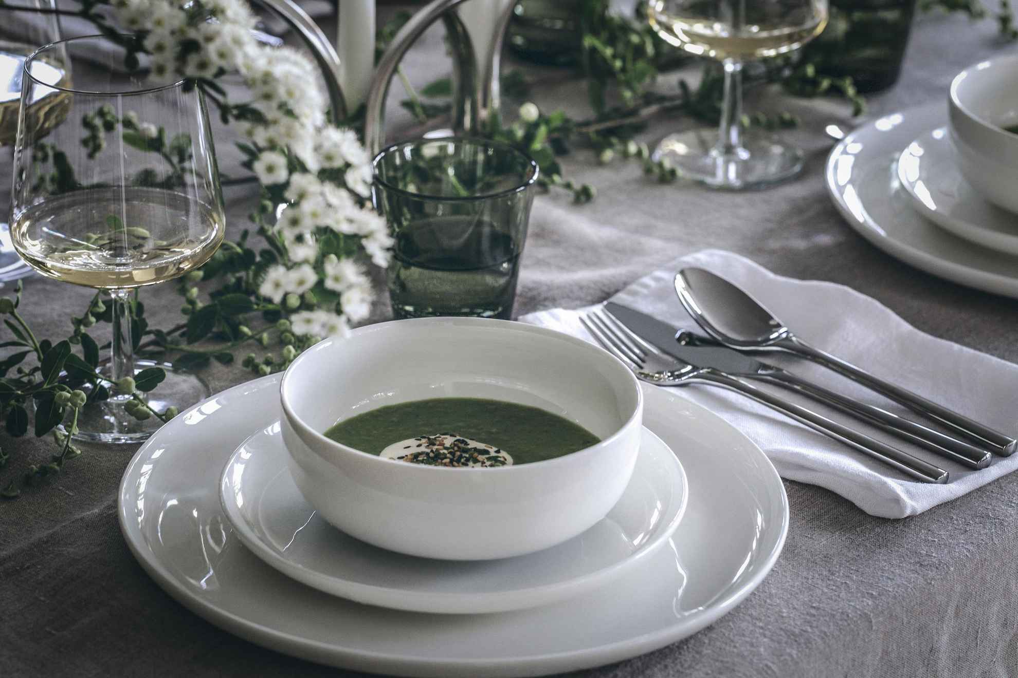 Gros plan sur une table où sont disposés des assiettes blanches et un bol de soupe aux pois verts, un verre à vin avec du vin blanc. Des fleurs blanches décorent la table.