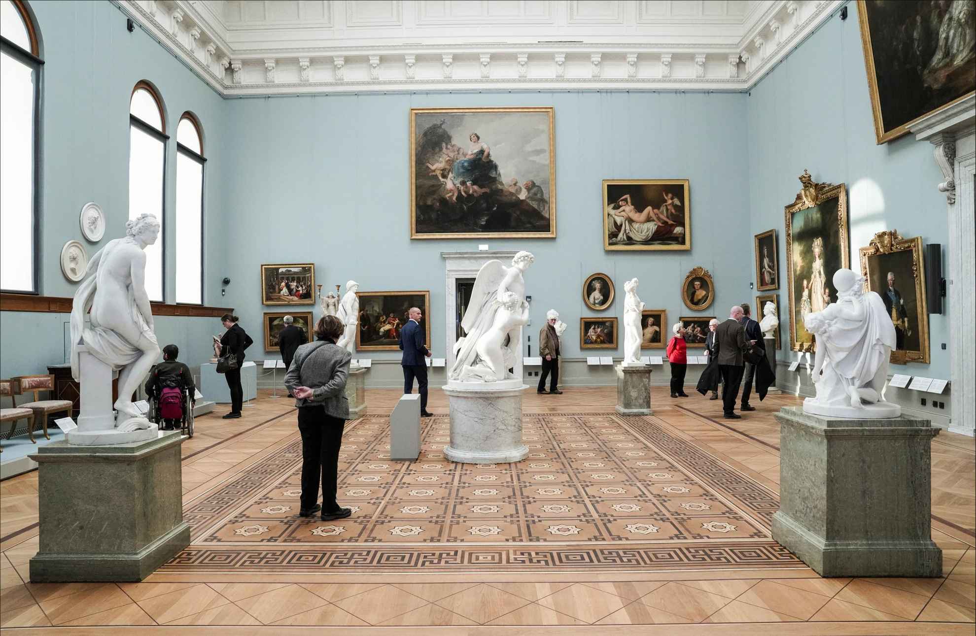 Des personnes se promènent dans une grande salle où sont exposées des peintures et des sculptures.