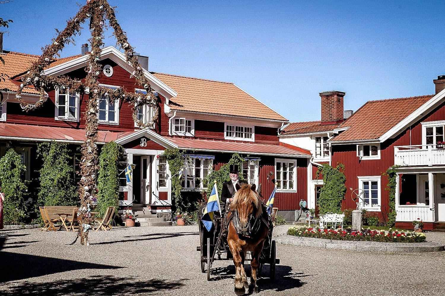 Une grande maison en bois rouge avec des bords blancs pendant l'été. Devant la maison, il y a un mât d'été typique de Midsommar et à côté un cheval avec une calèche et deux drapeaux suédois.