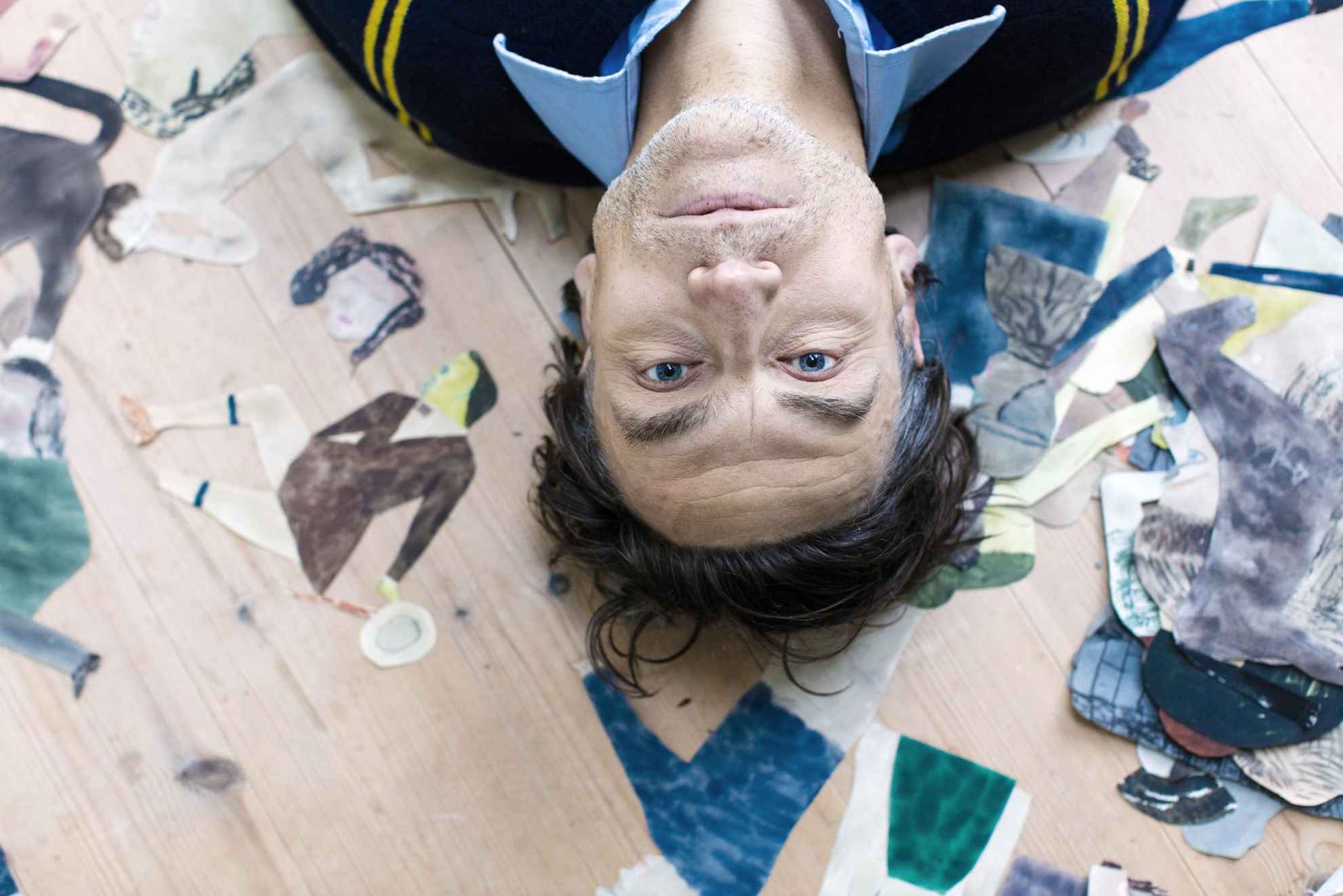 A close-up image taken from above at the artist Jockum Nordström when he is lying on the floor together with his paintings and illustrations.