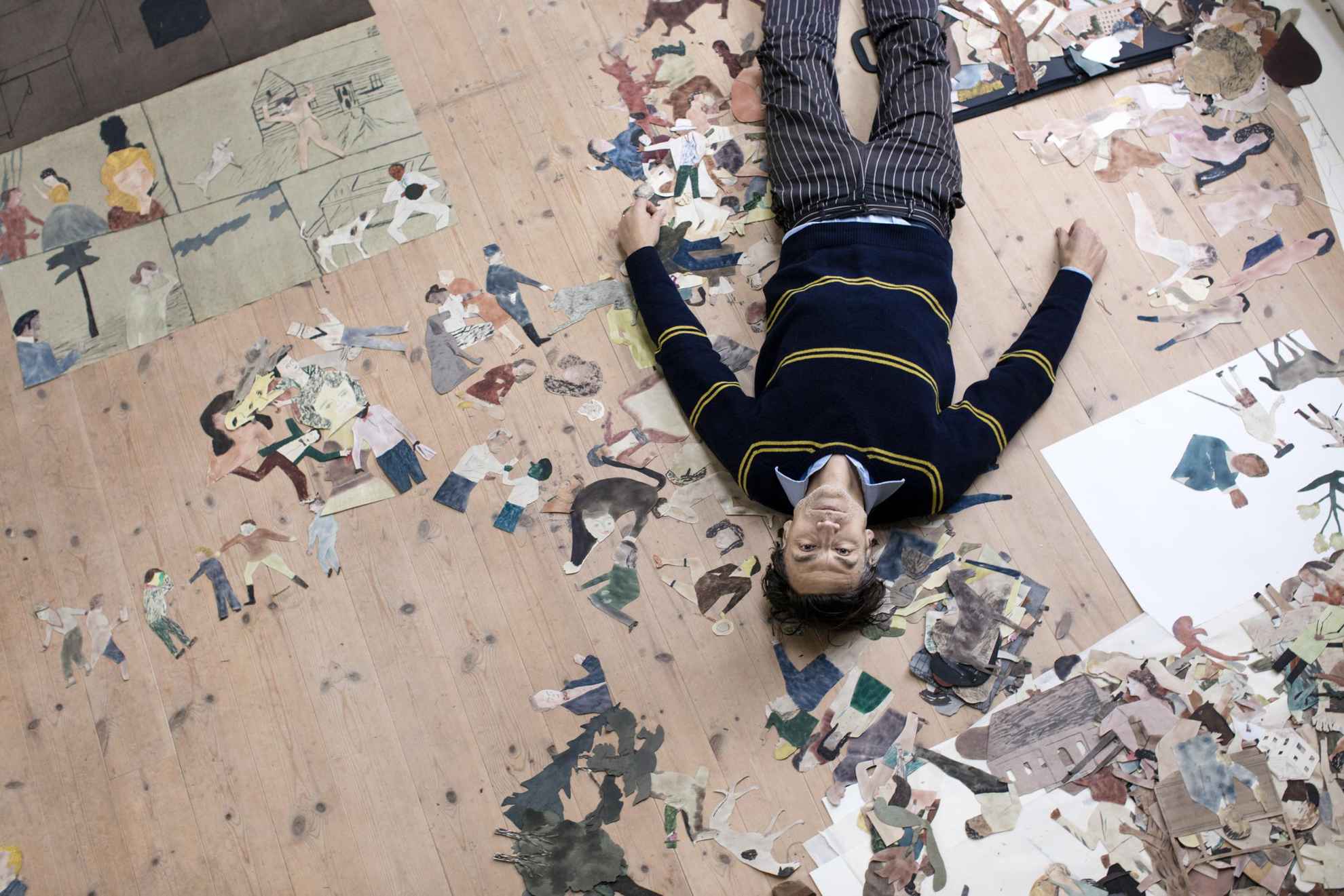 A image taken from above at the artist Jockum Nordström when he is lying on the floor together with his paintings and illustrations.