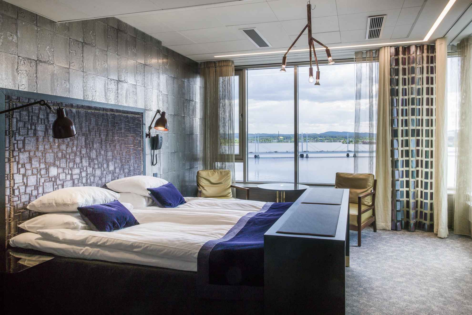 Une chambre d'hôtel avec vue sur un lac en été. La chambre possède un lit double, une petite table ronde et deux chaises.