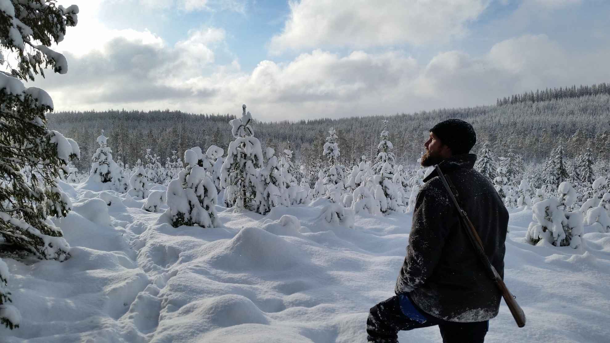 Un homme se tient debout dans une forêt enneigée avec un fusil de chasse accroché au dos.