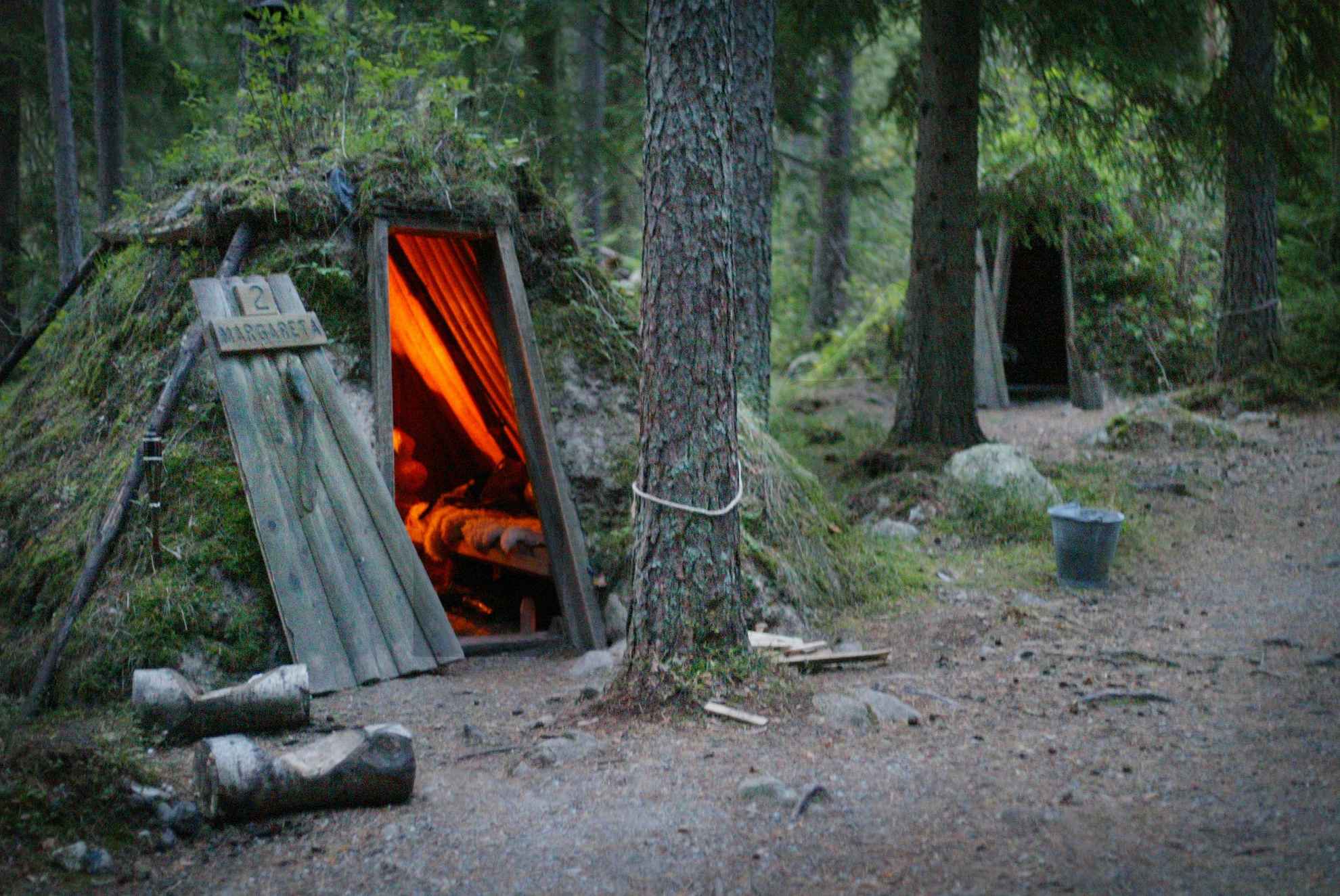 Deux huttes recouvertes de mousse sont situées dans une forêt. Il y a un feu de cheminée dans l'une des huttes.