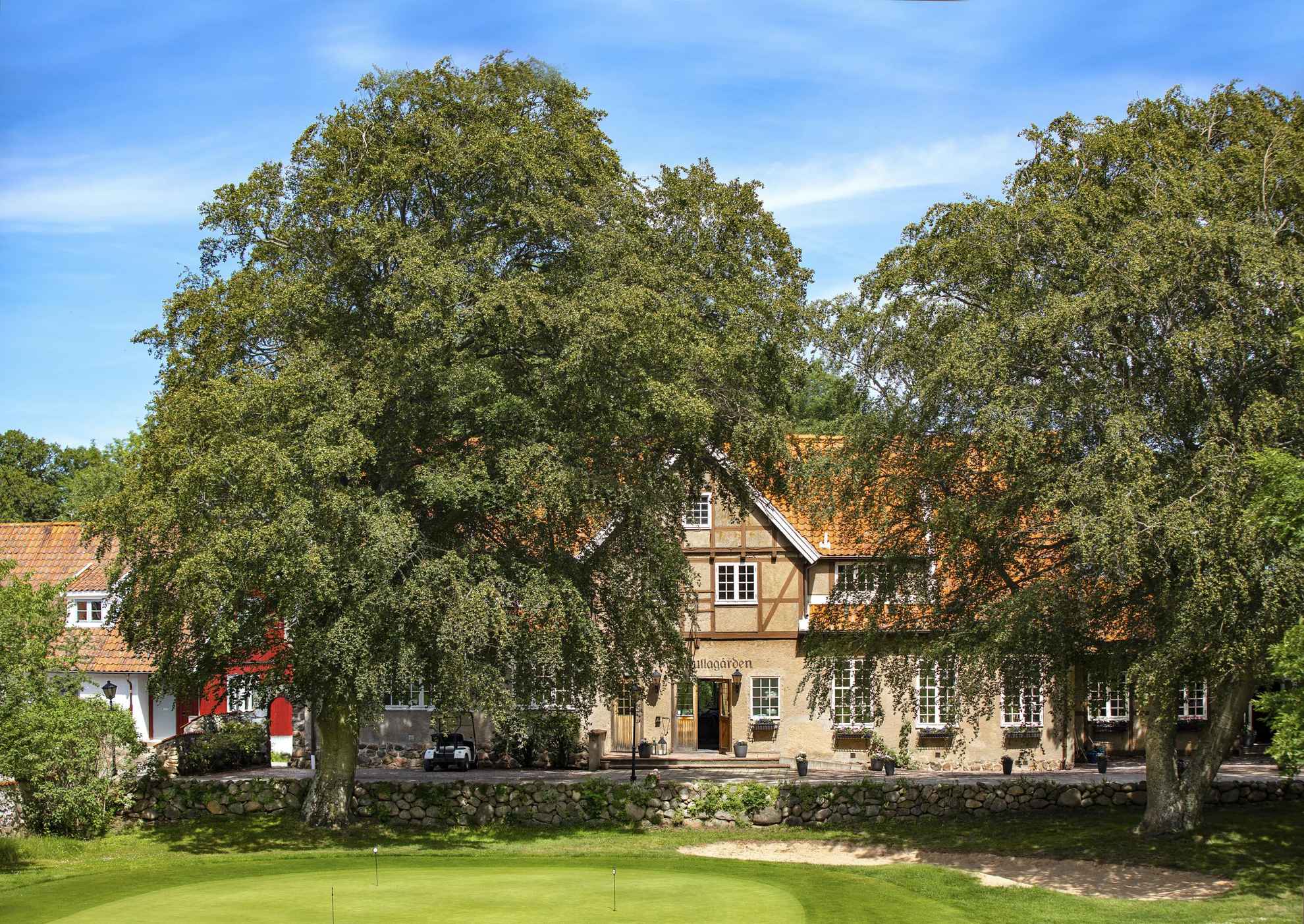 The lush exterior of Kullagårdens Wärdshus in Skåne. The red-and-brown building is surrounded by green trees and it is a sunny summer day.