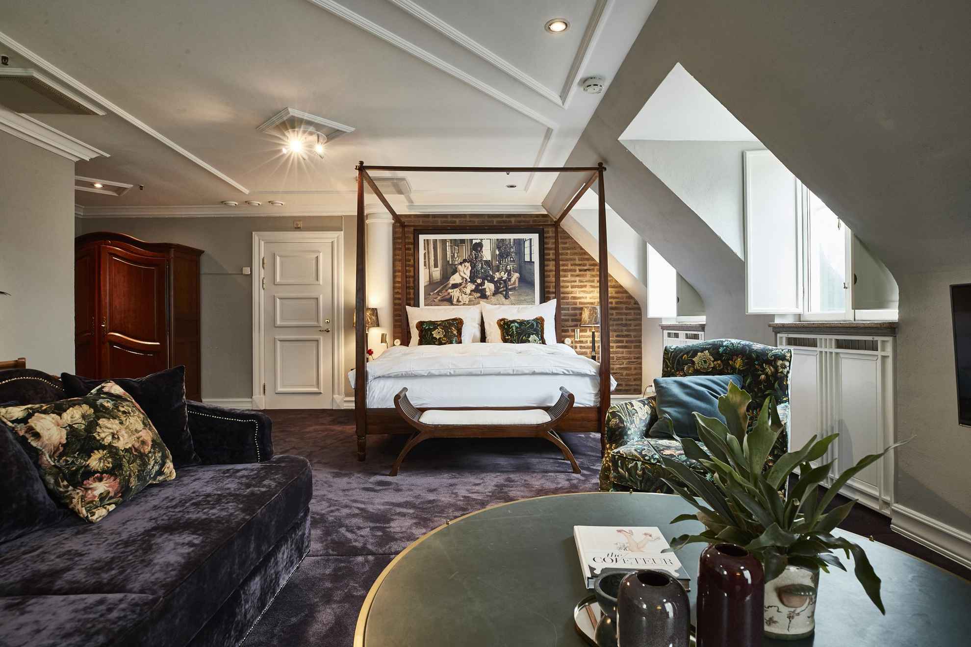 Une grande chambre d'hôtel avec de la moquette, un canapé en velours, une table basse verte, un lit double et une armoire en bois.