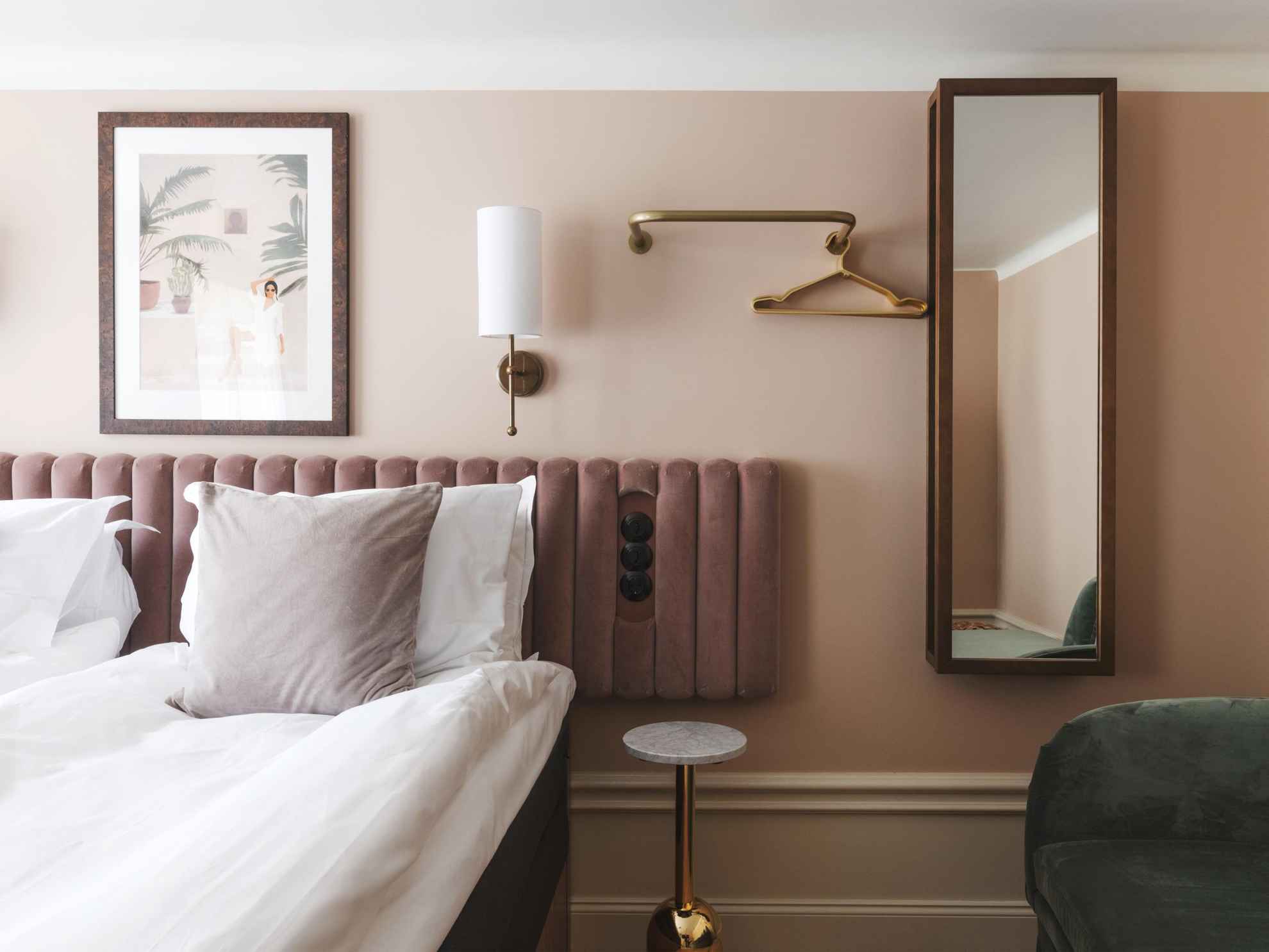 Une chambre d'hôtel avec des murs peints en beige et un plafond blanc. Sur les murs à côté du lit double, il y a une applique en laiton, un tableau, un miroir et un porte-manteau.