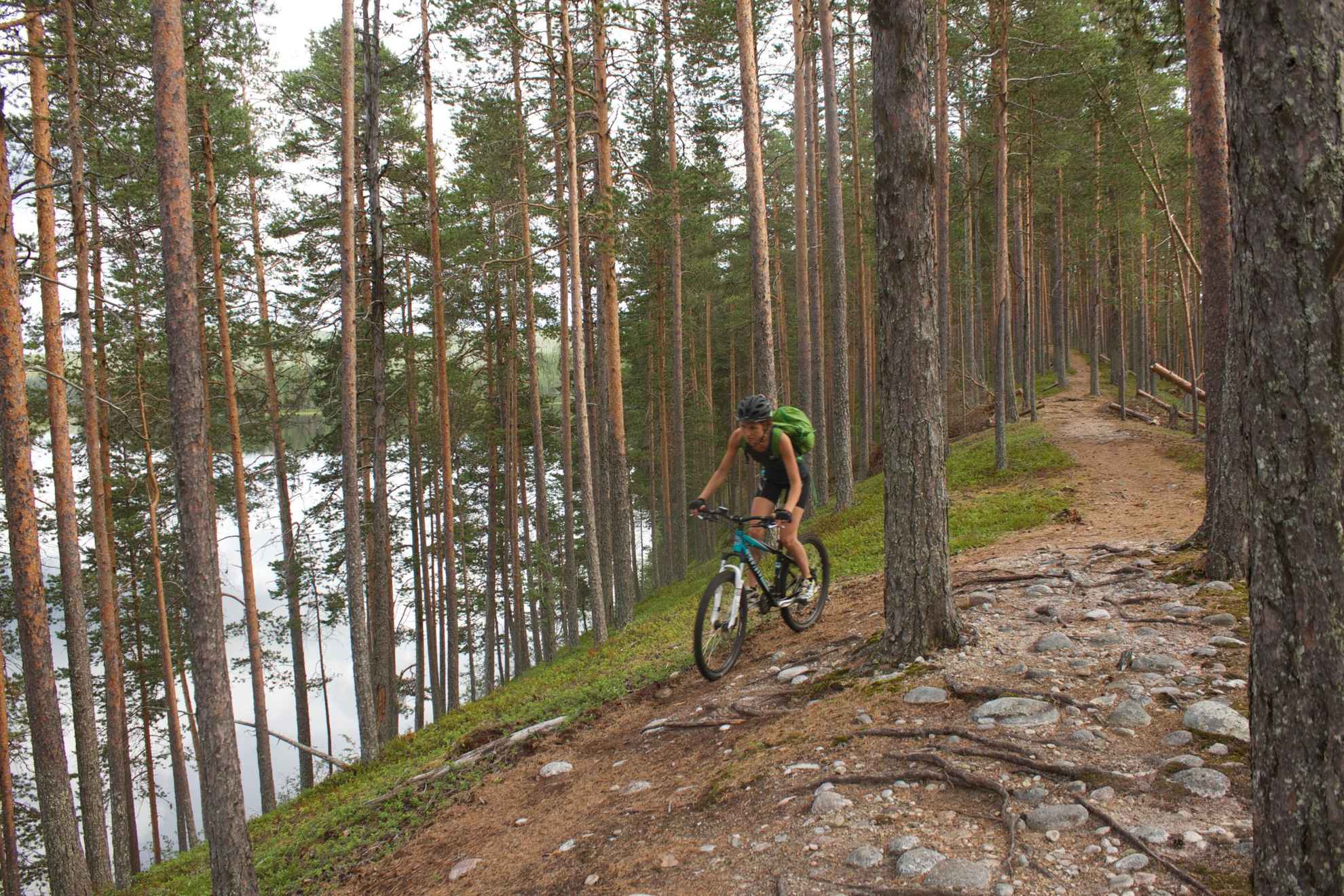 Une femme avec un sac à dos fait du vélo dans une pinède. Vous voyez un lac entre les arbres.