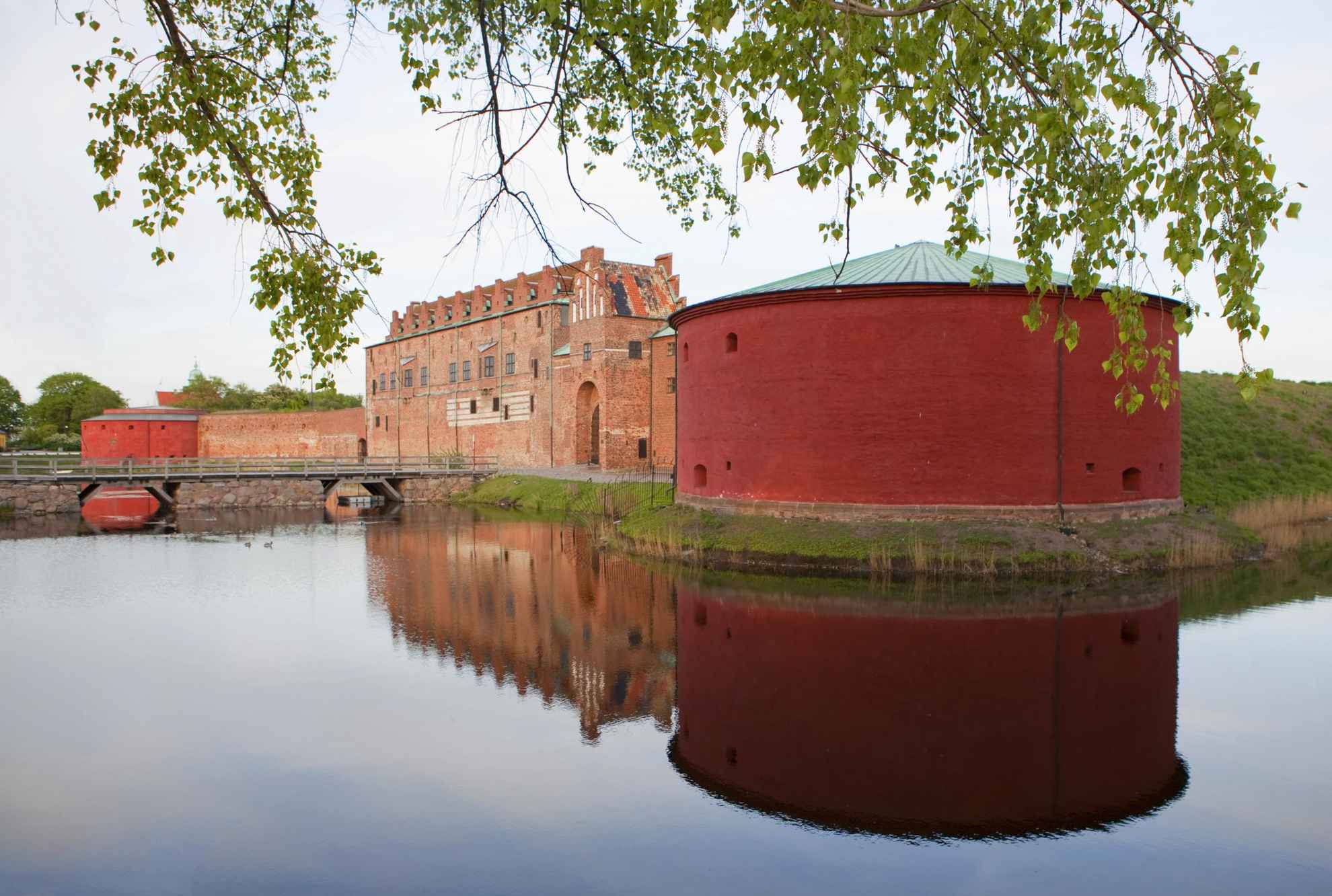 Le château de Malmöhus se compose d'une grande tour ronde avec à côté, un grand bâtiment en brique. Le château est entouré d'eau. En arrière-plan, on aperçoit une autre tour ronde de l'autre côté du bâtiment. Du feuillage se trouve au premier-plan.