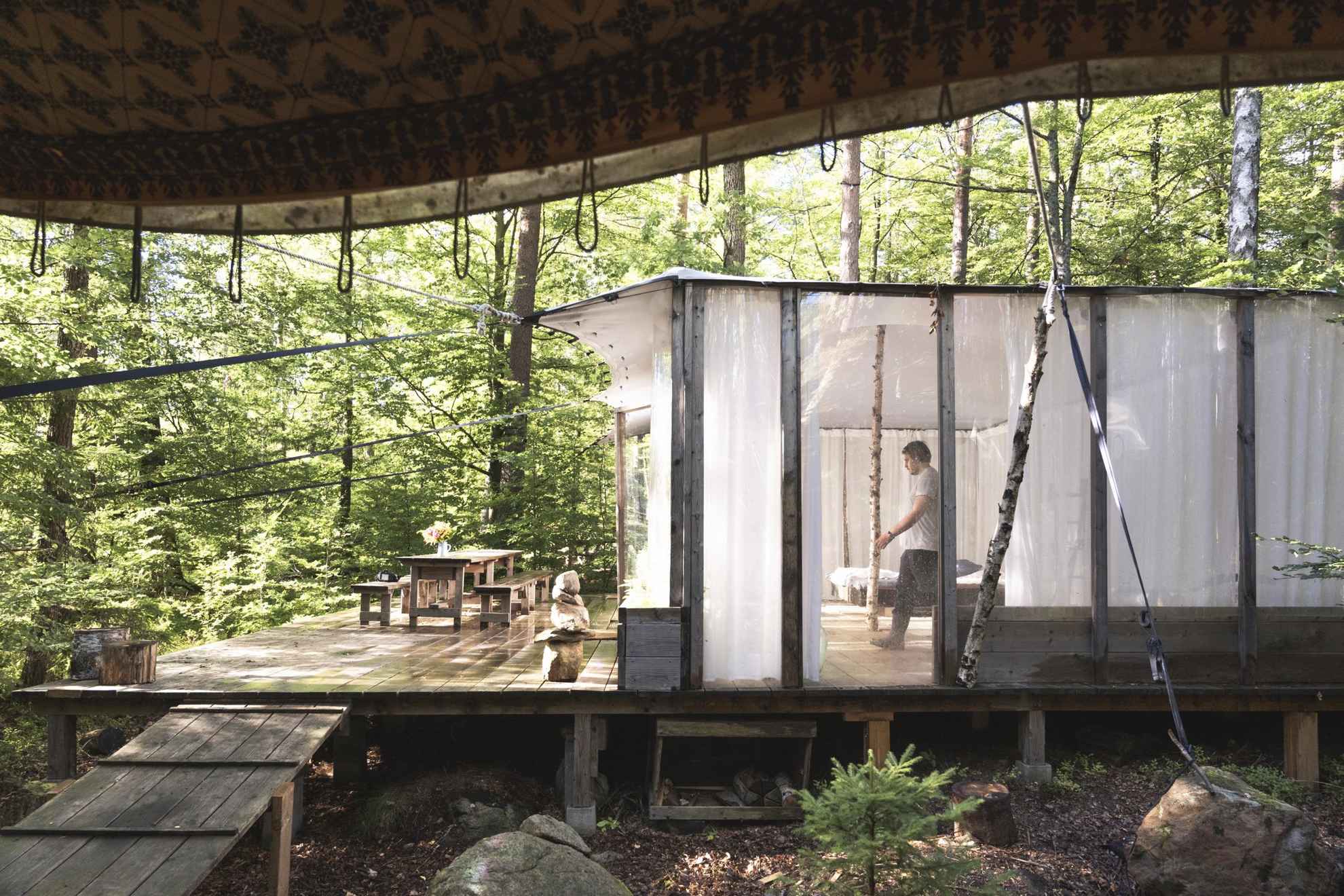 Vue extérieure sur une cabane légère et transparente dans la forêt, avec du feuillage vert tout autour et un homme marchant à l'intérieur de la cabane.