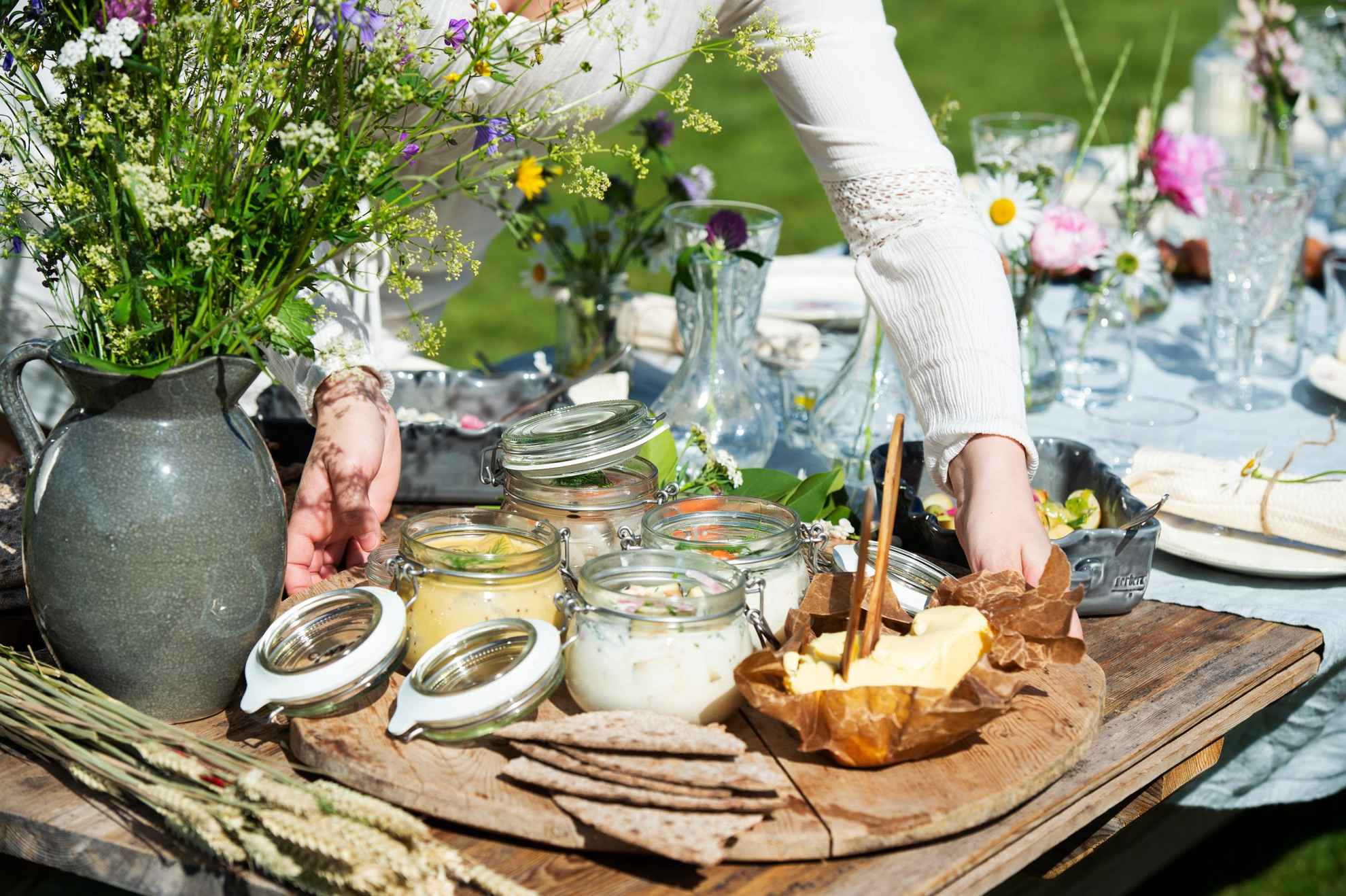 Une femme pose un plateau contenant des harengs marinés, du beurre et du pain sur une table préparée pour le déjeuner. À côté du plateau se trouve un vase avec des fleurs sauvages.