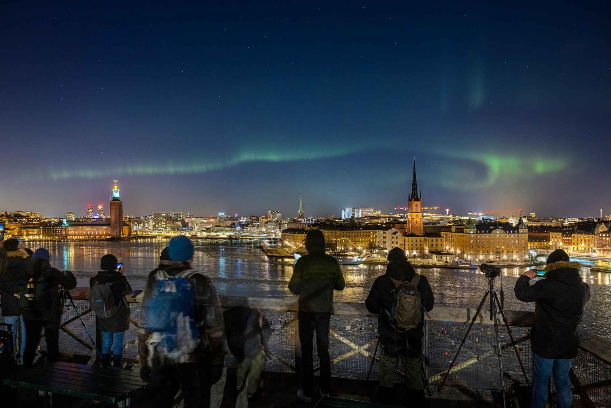 Une aurore boréale sur un paysage urbain, avec des photographes qui font la queue pour prendre une photo.